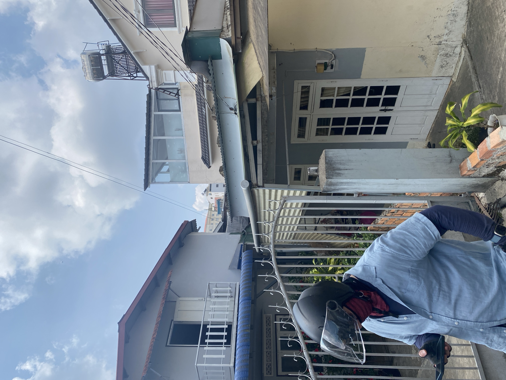A partially obstructed view of a person wearing a motorcycle helmet and a light blue shirt, standing behind a wrought-iron fence. Beyond the fence are buildings, including a house with white double doors and another with a blue and white facade. The sky above is partly cloudy. The location is presumed to be in or near Lâm Viên - Đà Lạt, Vietnam.