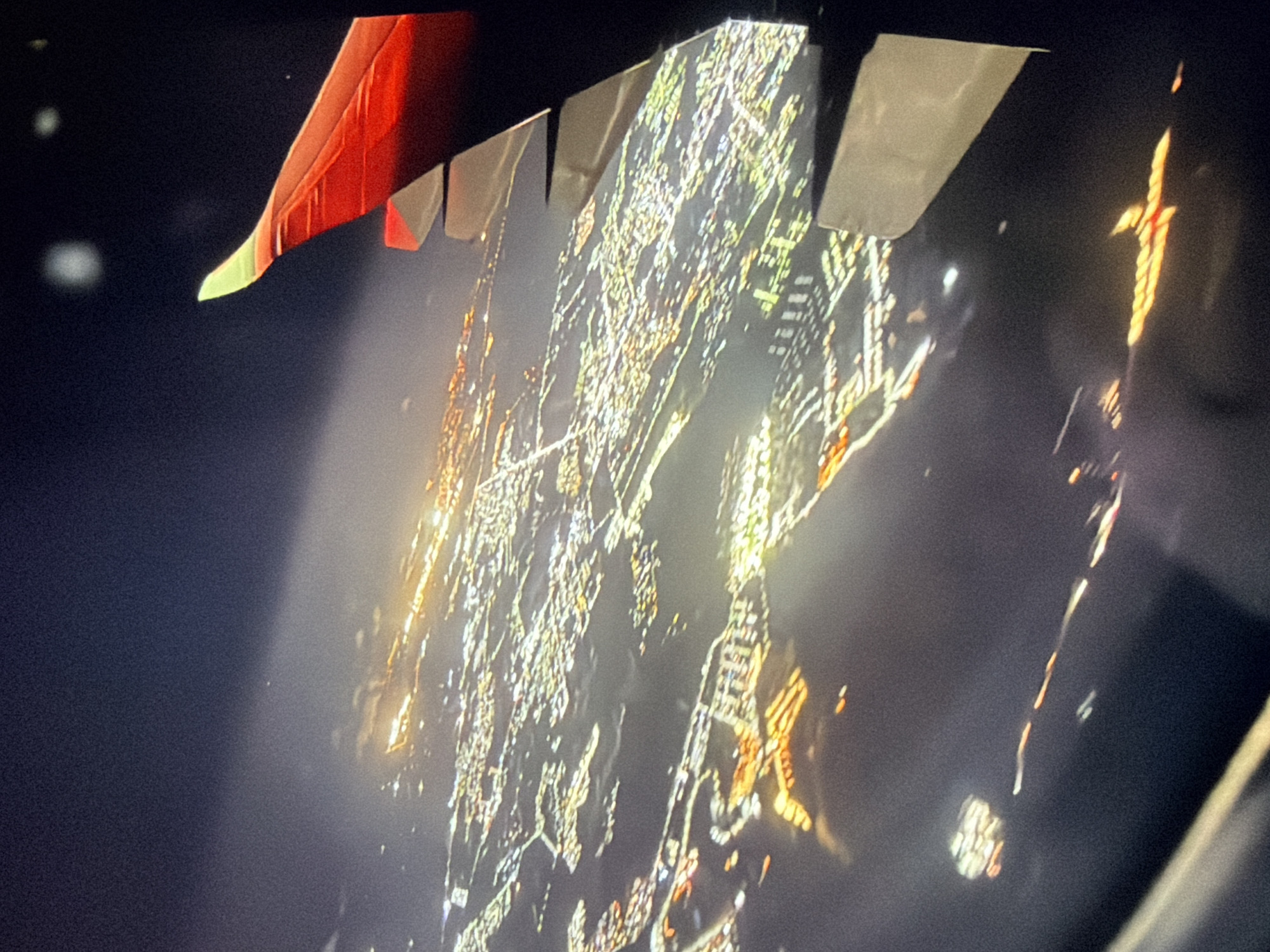 The image presents an aerial view of a sprawling city at night, illuminated by countless artificial lights, taken from an airplane. A prominent red airplane wing with a distinctive bright green tip is visible in the upper left corner, indicating the vantage point. Several light-colored, angular structures, likely part of the aircraft's wing or fuselage components, extend from the top edge of the frame.
The city below is characterized by a dense, intricate network of bright yellow, orange, and white lights that form clear grid-like patterns, suggesting streets and urban development. A particularly notable feature among the city lights is a distinct, bright, cross-shaped light formation visible in the upper right portion of the illuminated area. The background is a dark, starless sky, with some faint, blurry lights sparsely distributed in the distance. The scene captures the experience of flying over a populated urban area after dark. No visible text is present in the image.