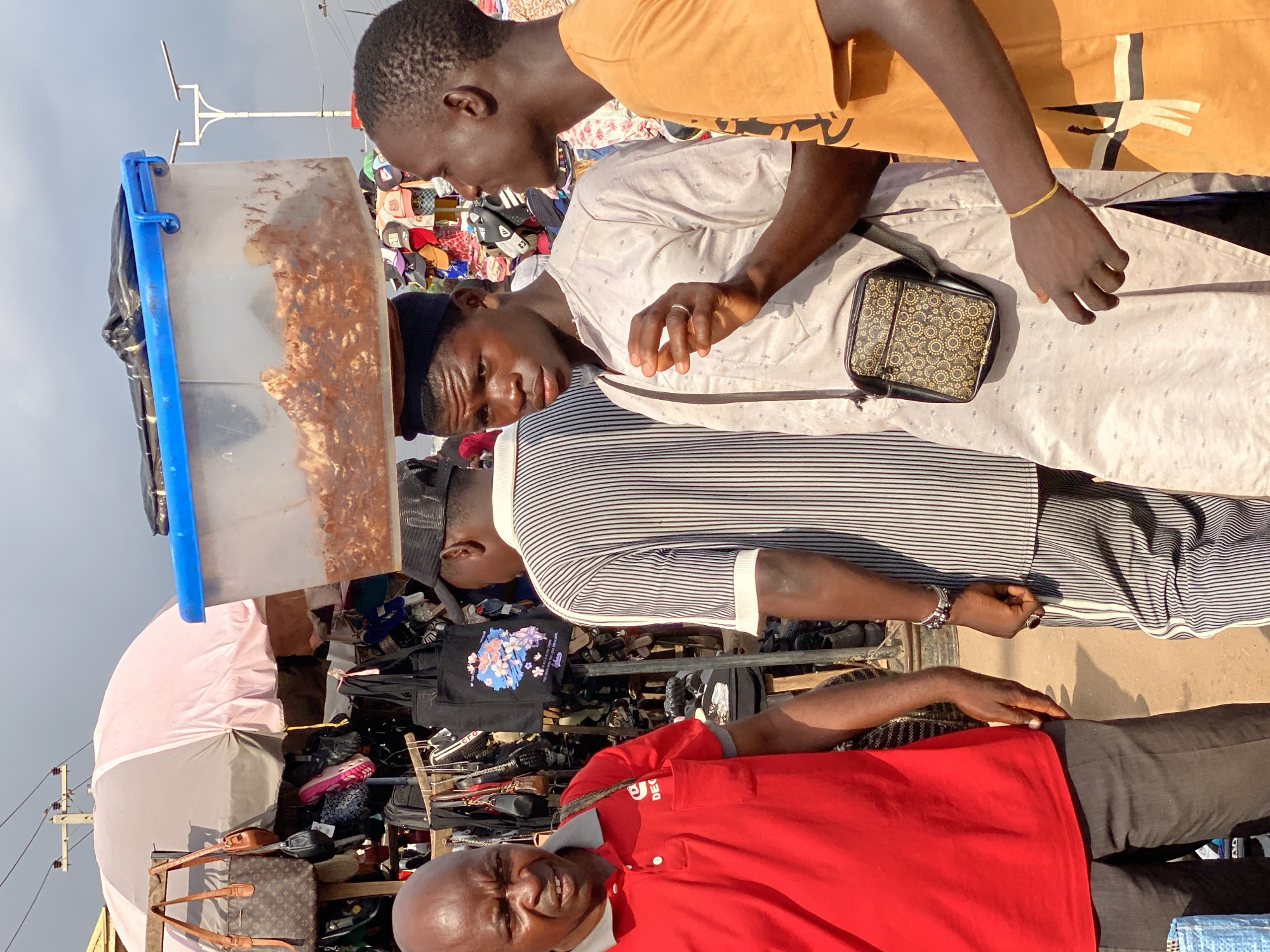 The image shows a group of people at an outdoor market in Sabon Tasha, Nigeria. In the foreground, an older man in a red polo shirt and black pants looks at the camera with a slight smile. Behind him, a younger man in a striped shirt and black pants is partially visible. To the left, a clear plastic container with some items inside is propped up by a blue handle. In the background, several people are visible, including one in an orange t-shirt and another in a white patterned shirt with a black bag slung across his chest. A variety of goods, including hats and bags, are displayed on racks and tables. The sky is visible and appears bright, suggesting it is daytime. The overall scene depicts a bustling market environment with people engaged in shopping or selling.