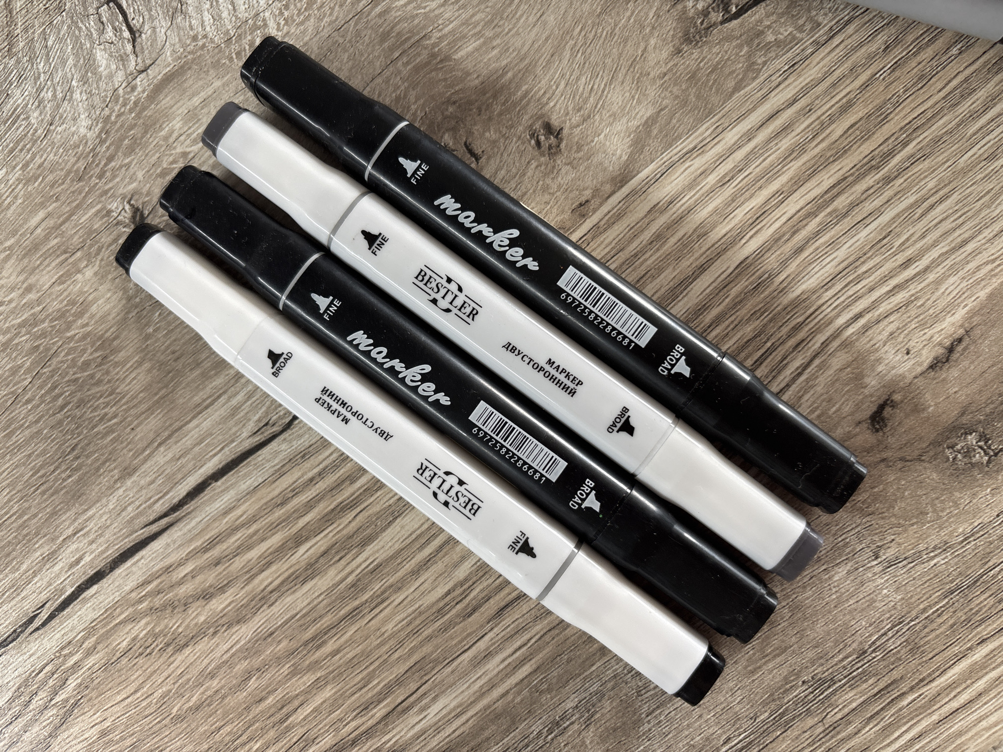 Four markers, two white and two black, are arranged diagonally on a wooden surface. The markers are branded "BESTLER" and have "BROAD" and "FINE" labels, indicating they are double-sided. The wooden surface has a natural grain pattern and appears to be a table or floor. The scene is well-lit, with shadows cast by the markers. There are no people or animals visible in the image. The location is not specified in the image.