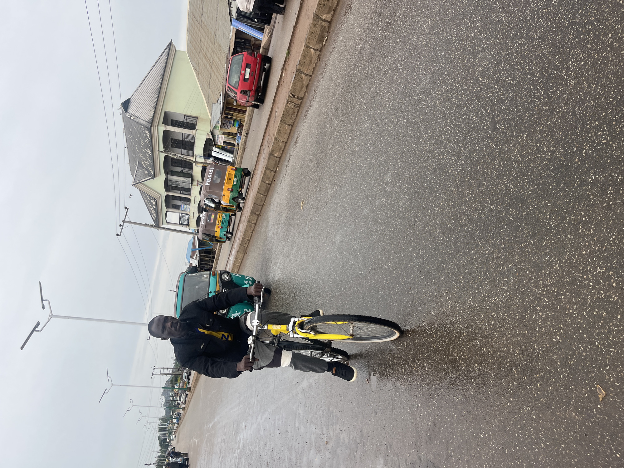A man is riding a yellow bicycle on a wet road in Bukuru, Nigeria. The man is wearing a black jacket over a yellow shirt and grey pants. He is looking towards the camera. In the background, there are several auto-rickshaws, a red car, and a building with a green facade and a dark roof. The sky is overcast. The road appears to be asphalt and is wet, indicating recent rain. Streetlights line the road.