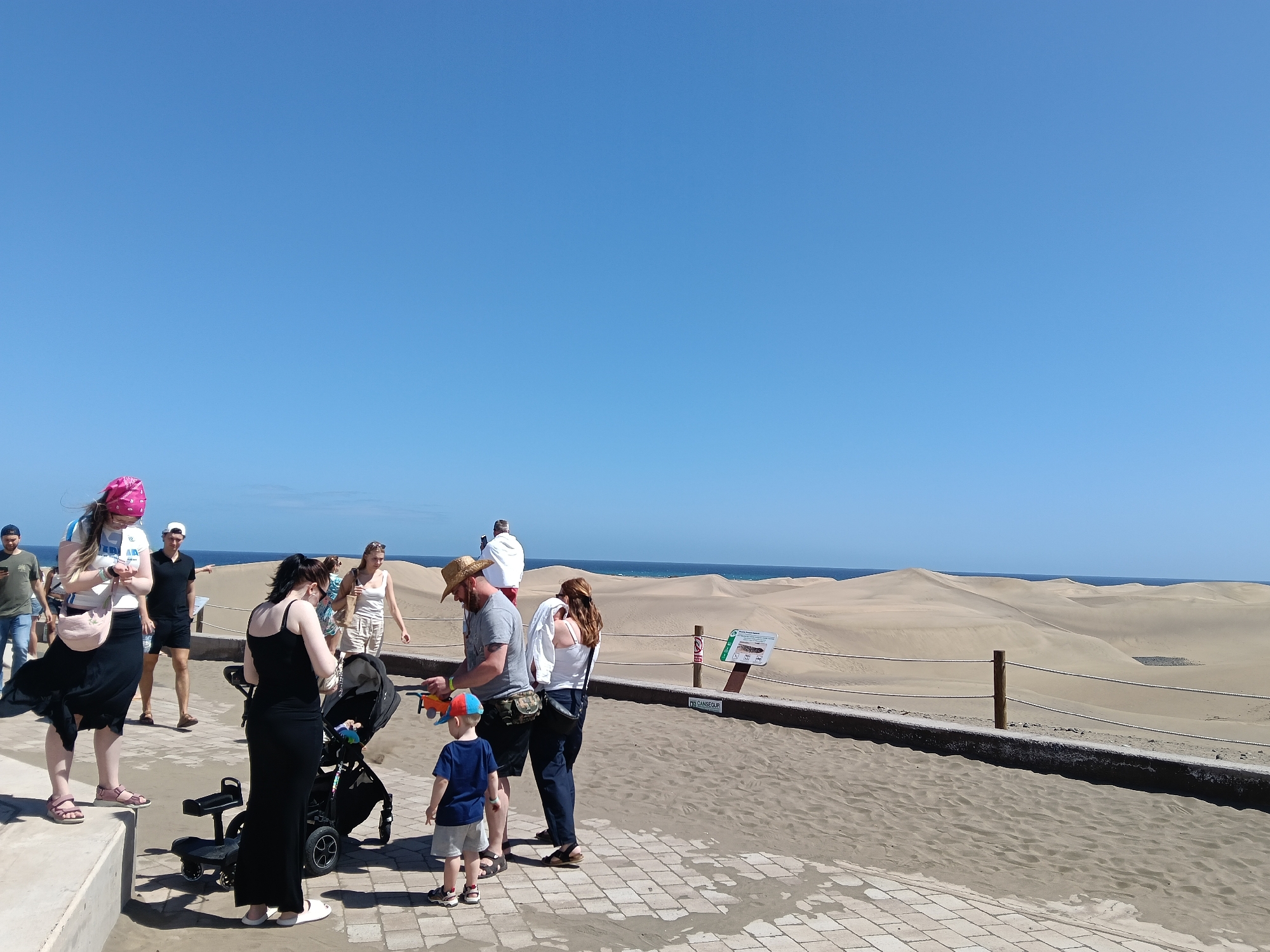 The image shows a group of tourists standing on a tiled walkway at the edge of the Maspalomas Dunes in San Bartolomé de Tirajana, Spain. The sky is clear and blue, and the sea is visible in the background, beyond the dunes.
The tourists consist of people of all ages including children. A woman with a pink head covering is looking at her phone. Another woman in a black dress stands next to a stroller. A man in a straw hat is standing next to a child wearing a blue shirt and a red and blue cap. There is a low wall separating the walkway from the sand dunes. A sign with information about the dunes is visible.