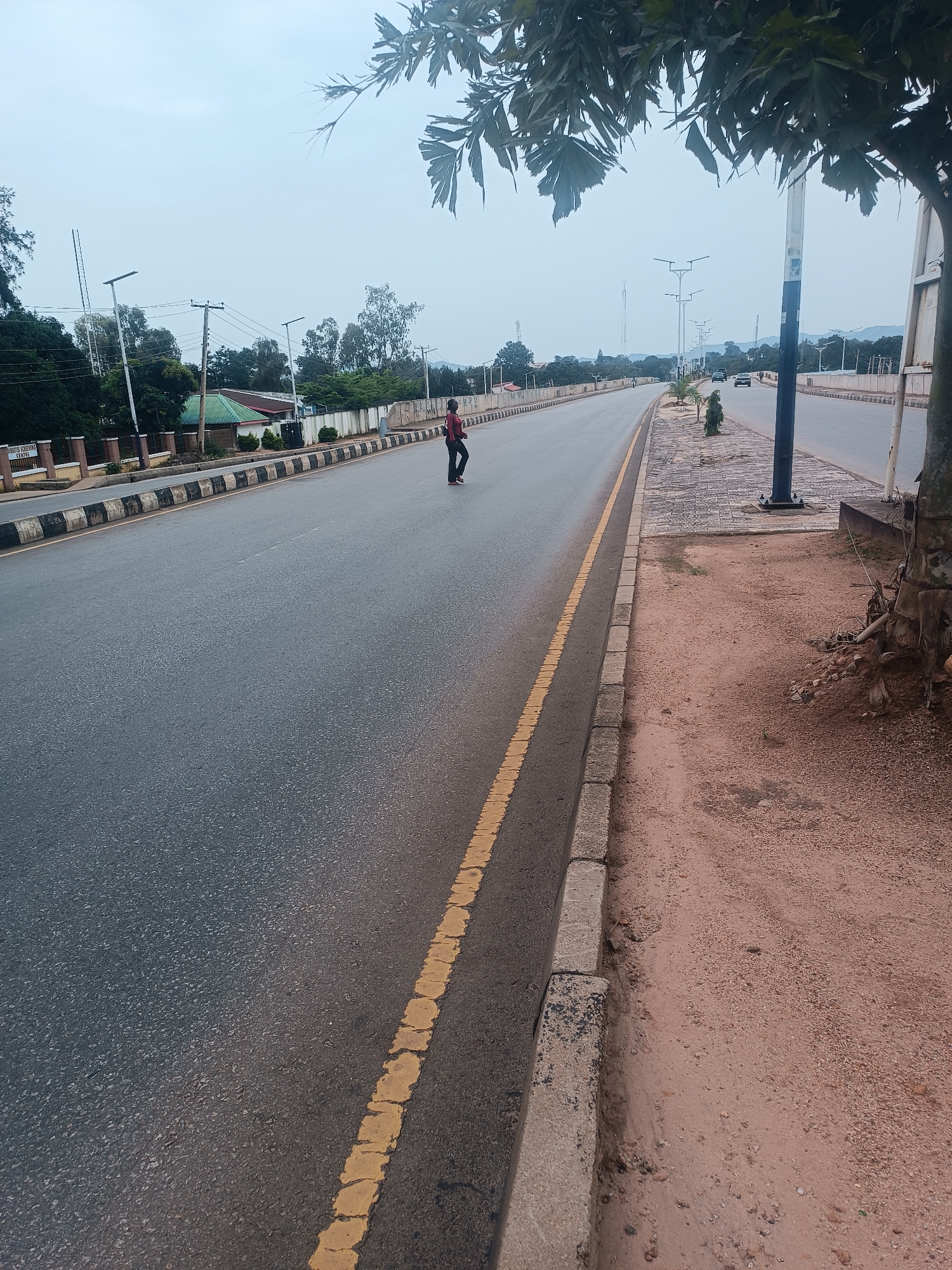 The image shows a wide asphalt road in Jos, Nigeria, with a person in dark clothing standing on the road, facing to the left. The road has a yellow dividing line and a black and white striped curb. On the left side of the road, there are buildings, trees, and streetlights. The sky is overcast. The ground on the right side of the road is sandy. The image appears to be taken from a slightly elevated position, looking down the road.