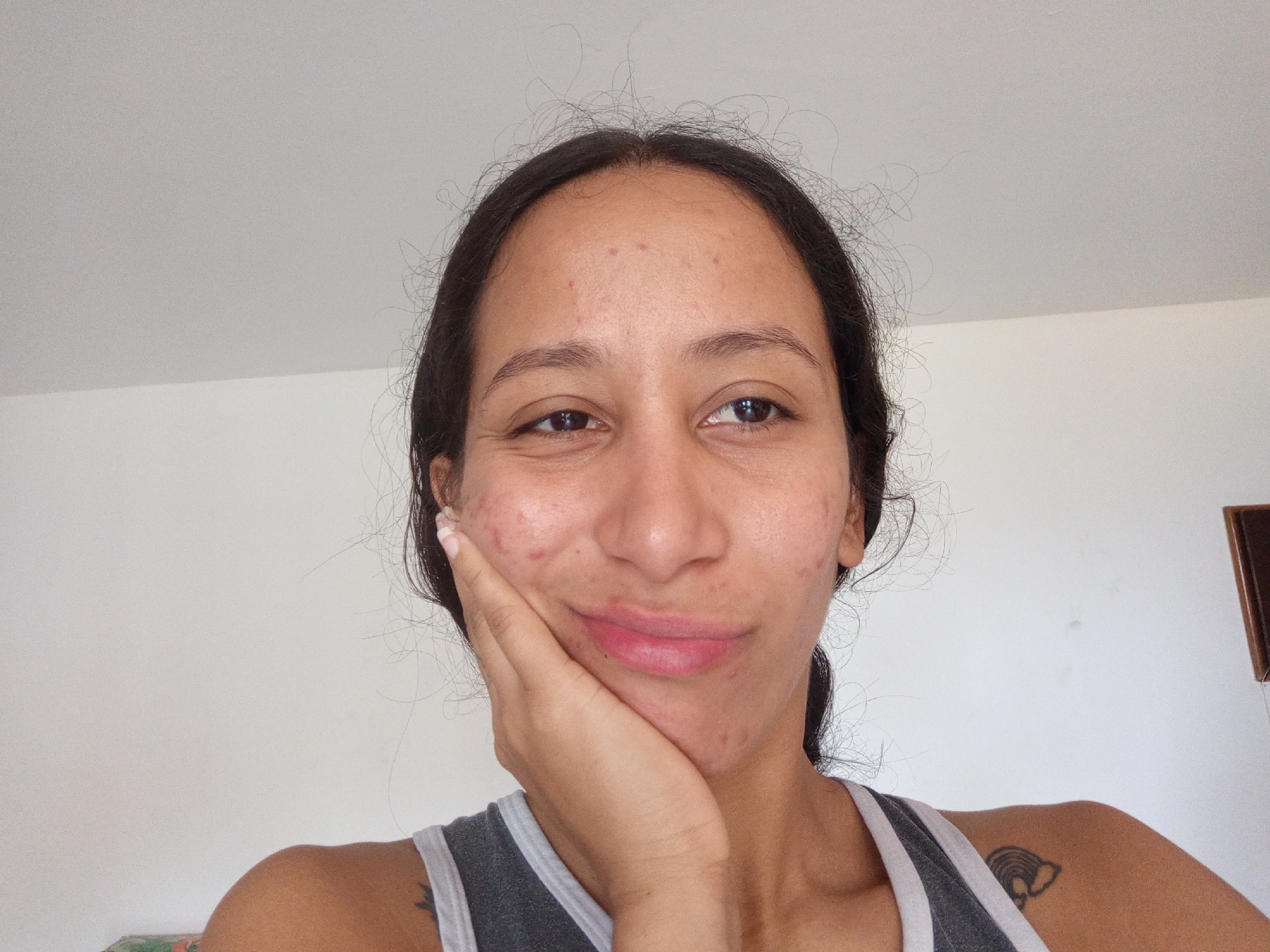 A head and shoulders shot of a woman with dark hair pulled back from her face, smiling slightly. She is wearing a gray tank top. She has her right hand on her cheek. There is a tattoo on her left shoulder. The background is a white wall. It is impossible to determine the location in Porlamar, Venezuela based on the image alone.
