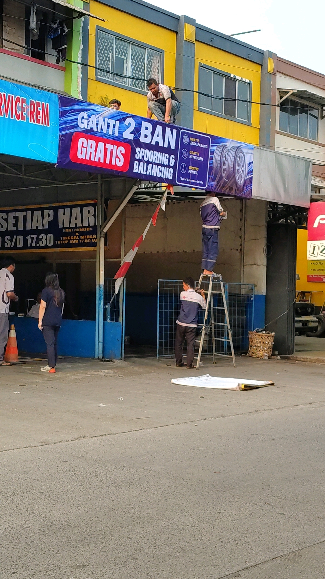 In Bencongan, Indonesia, a group of men are seen working on a banner advertising tire services. A man in a grey shirt and dark pants is crouched on the banner, holding it in place. Another man, wearing a blue uniform, stands on a ladder on the right side of the image, assisting with the banner installation. Below him, another man in a similar uniform is on the ground, holding the ladder steady. A third man in a blue uniform is also on the ladder, seemingly adjusting the banner from above. A large banner, predominantly blue with white and red text, is being affixed to the building. The text on the banner reads "GANTI 2 BAN GRATIS SPOORING & BALANCING". To the left, another banner with blue and white lettering states "SERVICE REM". The building itself is painted yellow with grey accents. In the foreground, a white rolled-up material is on the asphalt road, possibly another banner or protective covering. Orange cones are visible to the left of the frame, and a portion of a yellow truck is seen on the right. The scene suggests an outdoor commercial setting, likely a tire shop or automotive service center.