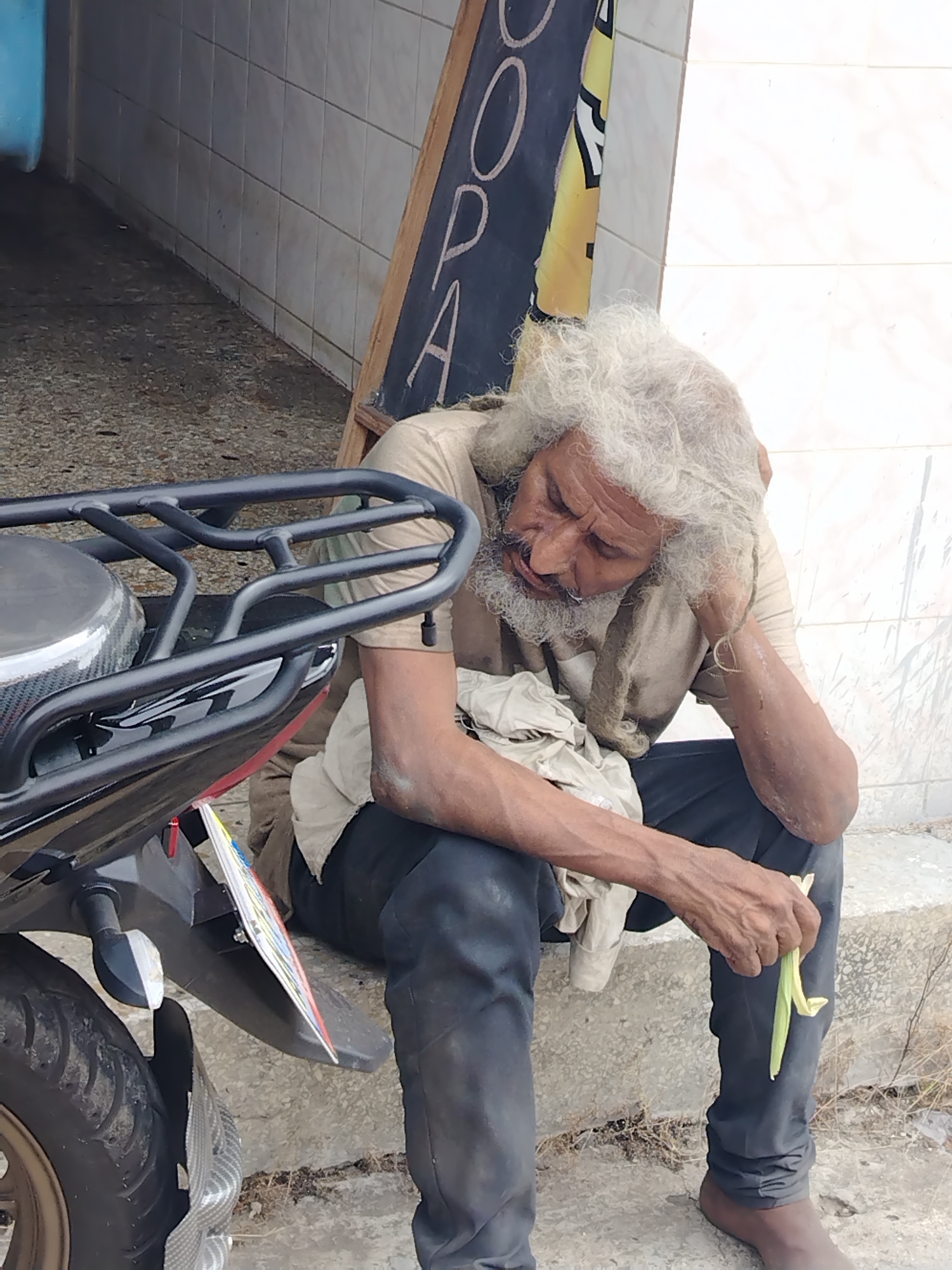 The image captures an elderly man resting outdoors in what appears to be an urban setting in Tinaquillo, Venezuela. He is seated on a concrete step or curb, leaning his head on his right arm, which is propped against his knee. His eyes are closed or cast downwards, conveying a sense of deep weariness or sleep.
The man has long, dishevelled white hair and a full white beard, and his skin shows signs of being weathered. He is wearing a light brown t-shirt and dark trousers, with a light-colored cloth draped over his lap. In his left hand, he holds a long, green, leafy object, possibly a corn husk or a similar plant part.
To the left of the man, the rear portion of a black motorcycle is visible, featuring a tire, a rear fender, and a black metal cargo rack. A colorful license plate is attached to the motorcycle's fender. The background includes a light-colored tiled wall on the right side of the frame and a textured, possibly concrete, ground surface. Partially visible against the tiled wall is a black chalkboard sign with white handwritten letters, displaying "O P A". The scene is illuminated by natural daylight.