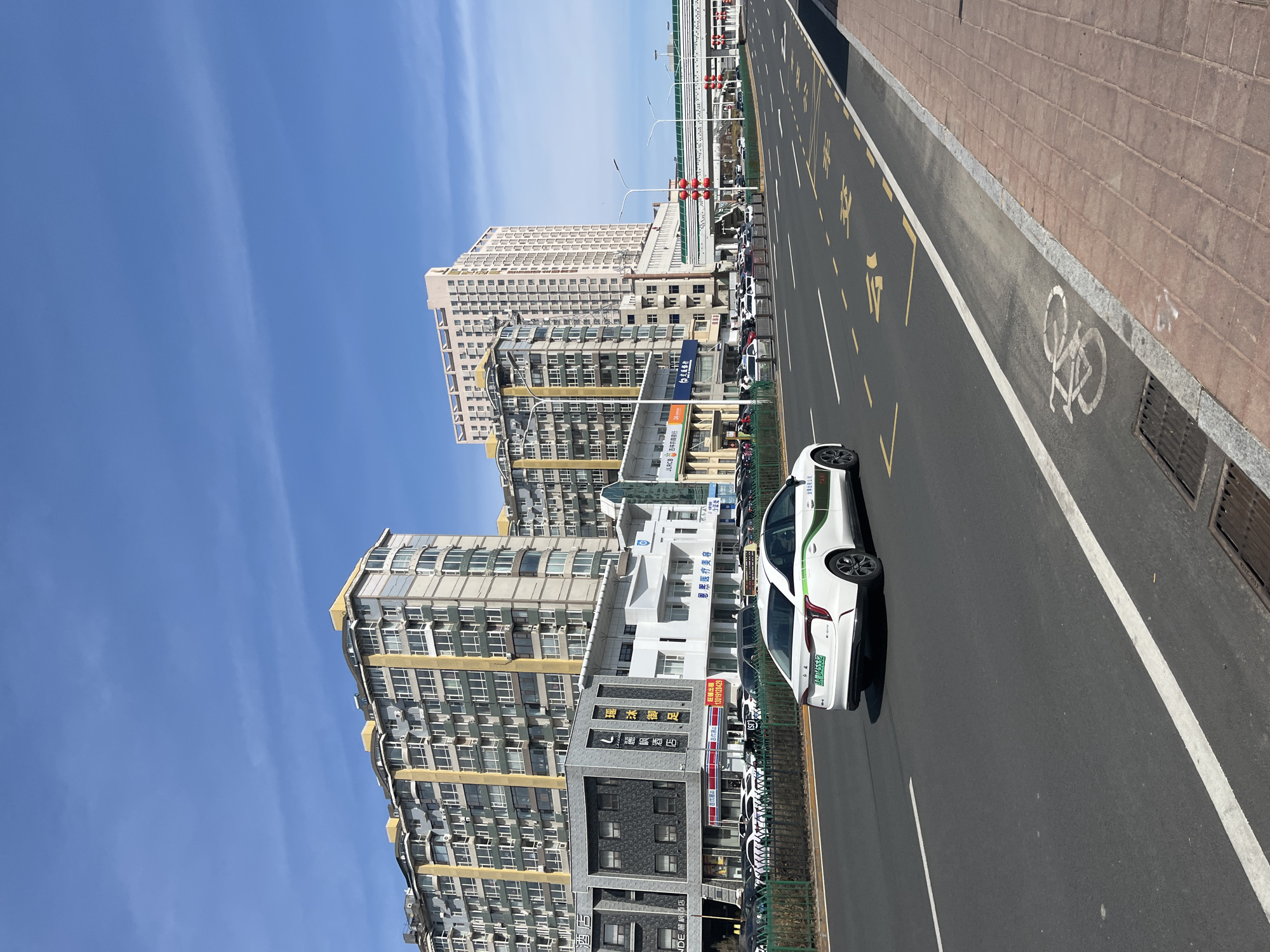 Here's a forensic description of the image:
**General Scene:** The image shows a street scene in what appears to be an urban environment, possibly Changchun City, China, based on the language of the visible signs. The perspective is from the side of the road, facing the city buildings. It appears to be daytime with clear blue skies.
**Buildings:** Several multi-story buildings with a mix of commercial and residential purposes line the street. These are characterized by repetitive window patterns, likely apartments. The architecture is modern, though somewhat utilitarian. Signage in Chinese characters is visible on many of the buildings, suggesting businesses are located there.
**Transportation:** A white car with green trim is moving on the road. The car has a license plate that reads "吉A D45532". The road has yellow painted lines and symbols, possibly indicating lane markings or speed limits. There is a dedicated bike lane with a bicycle symbol painted on the pavement.
**Street Details:** A brick paved area forms a sidewalk. There is a barrier or railing between the street and the buildings, and a row of parked cars alongside the curb.
**Signage:** The signage on the buildings includes names of various businesses such as hotels ("麗枫酒店"), medical clinics ("恩恩医疗美容"), and financial institutions ("吉林农農推行"). Phone numbers also appear on some of the signs.
