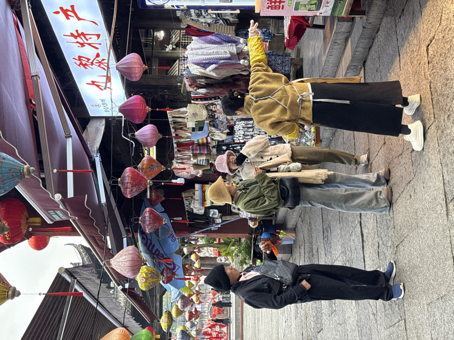 The image depicts an outdoor market scene in Pengshui Miao and Tujia Autonomous County, China, under an overcast sky. The street is paved with grey rectangular stone slabs.
In the foreground, four individuals are visible. From left to right:
1. A male individual wearing a black knit beanie, a black jacket over a light pink collared shirt, black trousers, and blue sneakers. He carries a dark grey patterned crossbody bag and holds a small orange object in his right hand. He is facing to his right.
2. A female individual wearing a light brown bucket hat, an olive-green jacket, light blue wide-leg jeans, and brown boots. She carries a dark crossbody bag and a light beige fringed scarf. Her head is tilted downwards, and she appears to be looking at a smartphone held in her hands. She is positioned slightly to the right of the male.
3. A female individual wearing a light pink bucket hat, a light beige fluffy jacket, brown trousers, and white sneakers. She is standing next to the second female, facing towards her left, with her mouth slightly open, suggesting speech or a reaction.
4. A female individual, seen from behind, wearing a mustard yellow jacket with white ties (possibly an apron), a black wide-leg skirt or trousers, and white chunky sneakers. Her dark hair is pulled back. She wears yellow sleeves or gloves on her forearms. Her right arm is extended horizontally to the right, in a pointing or gesturing motion. She is positioned facing away from the viewer, addressing the group in front of her.
The background features numerous vibrant, decorative paper lanterns of various shapes and colors (red, pink, orange, yellow, green, blue) suspended across the street above market stalls. Two stalls are visible with dark red awnings. The stall on the right has a prominent illuminated sign displaying red Chinese characters, "丸黎特产" (Wán Lí Tèchǎn), which translates to "Wanli Specialty Products." This stall displays clothing items, including knitted sweaters and scarves, along with bags and other merchandise. A table in the lower right foreground displays a white and green sign with the Chinese character "鲜" (Xiān, meaning fresh). Traditional Chinese architectural elements with dark, curved roofs are partially visible behind the stalls. A blue umbrella is visible under some lanterns near the left stall. Green potted plants are also discernible near the left side of the market area. Further down the street, blurred figures suggest more pedestrian traffic.