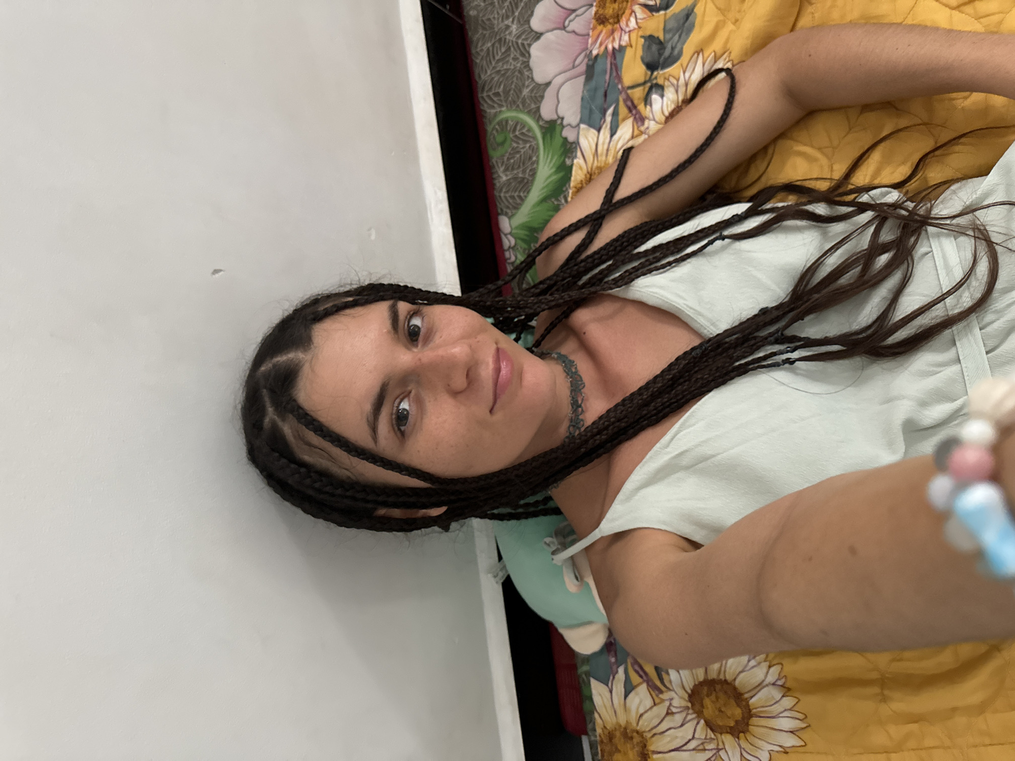 A woman with dark braided hair and a light blue dress is lying down on a bed with a floral yellow blanket. She is looking at the camera and smiling. The background is a white wall. The woman appears to be in Mũi Né, Vietnam, judging by the presence of a plush toy resembling a sea creature and a bedspread with a tropical flower pattern, commonly seen in tourist accommodations in the area.