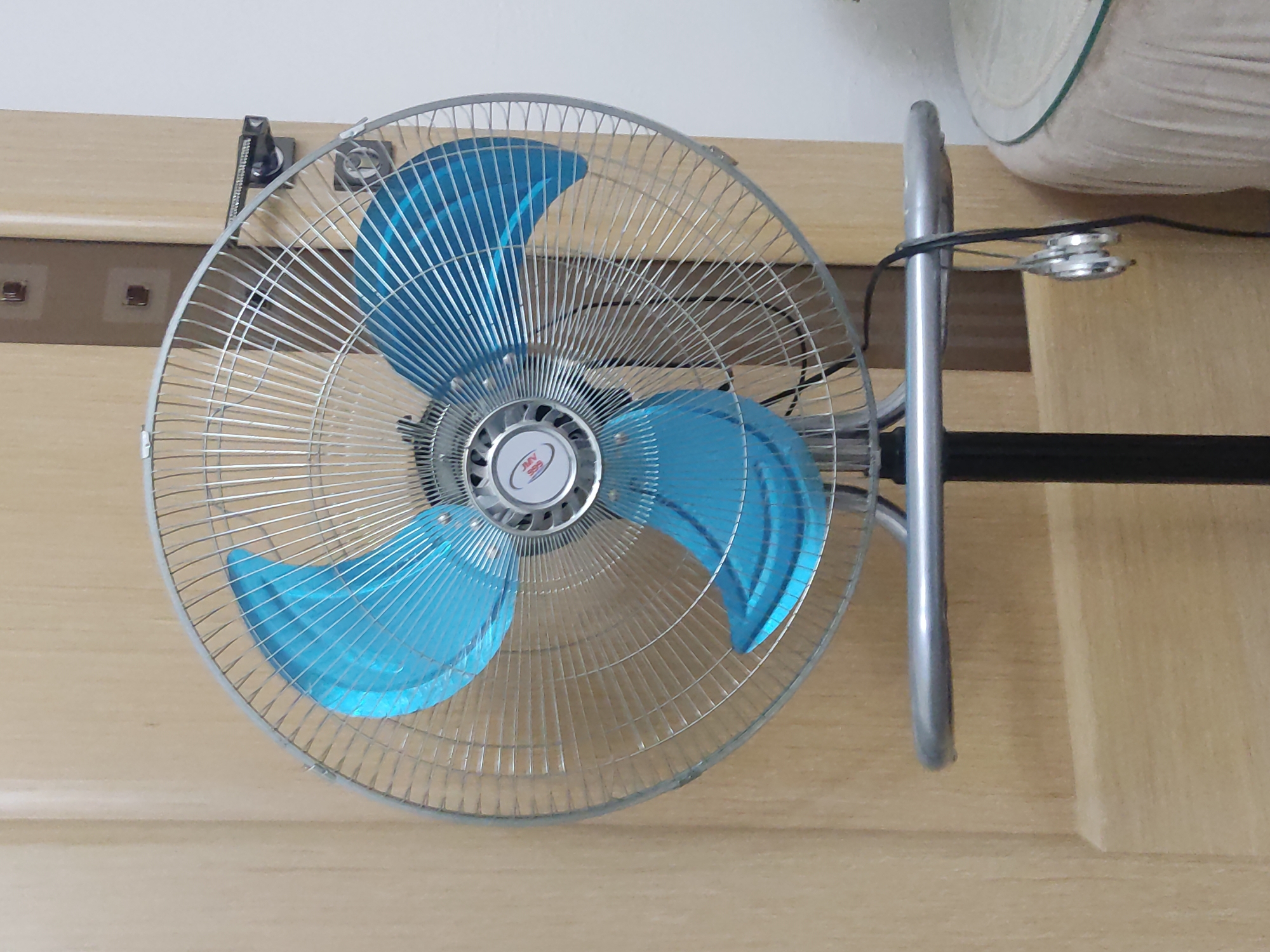 This is a medium shot, eye-level, indoor photograph of a blue-bladed electric fan with a metal grille. The fan is positioned in front of a light brown wooden door that has a vertical seam and two square decorative elements. The fan is mounted on a black stand, and a silver metal bar extends from the stand, supporting the fan. A black power cord hangs from the fan, with a pair of silver earbuds dangling from it, wrapped around the cord. To the right of the fan, a portion of a round table with a glass top and a beige tablecloth is visible. The lighting is even, and the overall scene appears to be an interior domestic setting. The city of Kurchaloy, Russia is not depicted in this image.