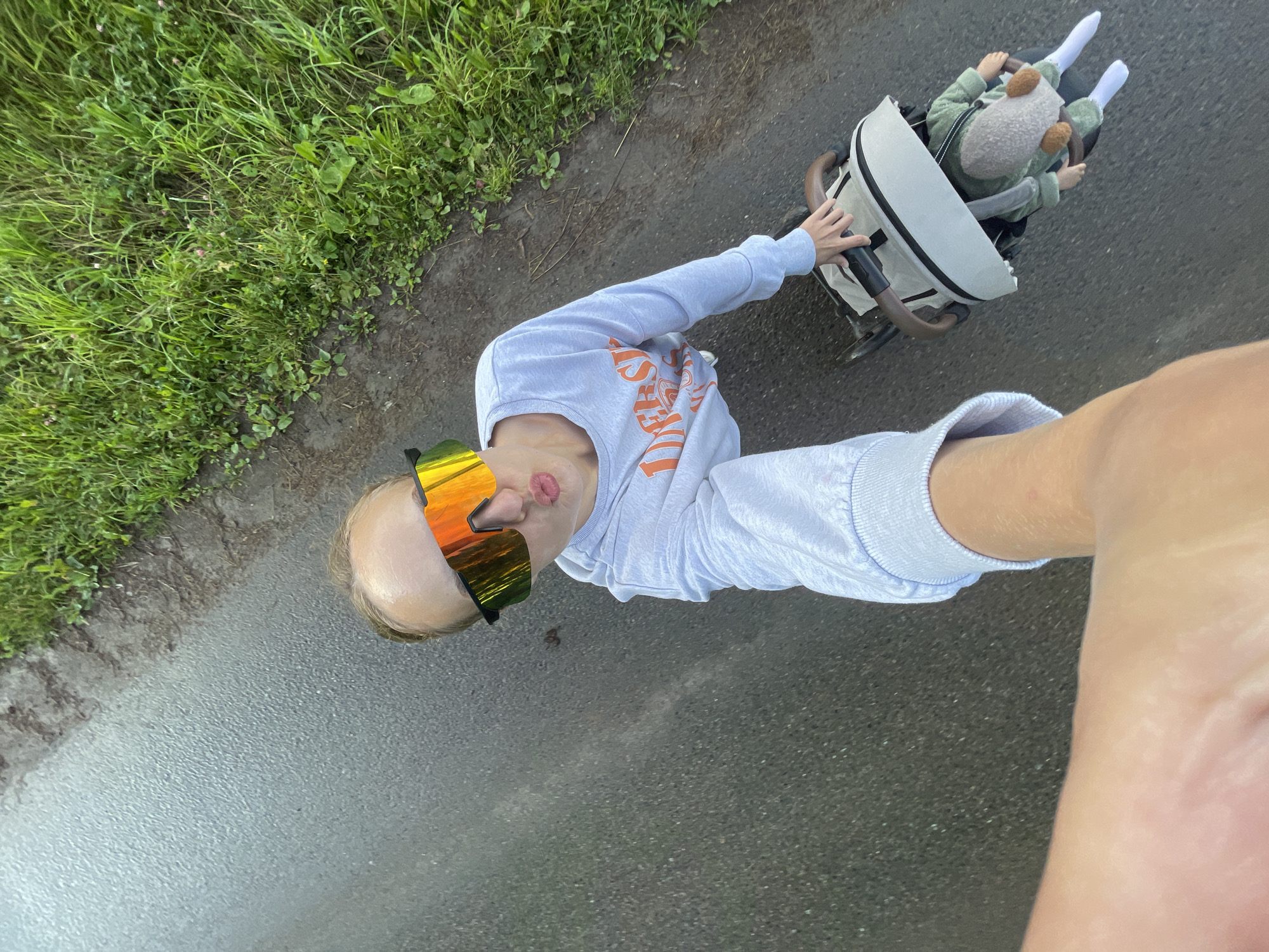 A person with blond hair pulled back is taking a selfie while wearing large, reflective orange and yellow sports sunglasses and a light blue sweatshirt with orange lettering. They are outdoors on a wet asphalt surface, possibly a road or path, with a grassy area to the right. In the lower right quadrant of the image, a stroller with a beige cover and brown handles is visible, containing a doll dressed in a green outfit and a grey hat with brown pom-poms. The person's left arm extends towards the camera, obscuring part of the frame, and their right hand is on the stroller's handle. The overall impression is a casual, outdoor scene, possibly in or around the village of Podshivalovo, Russia, given the presence of grass and a relatively unpaved road.