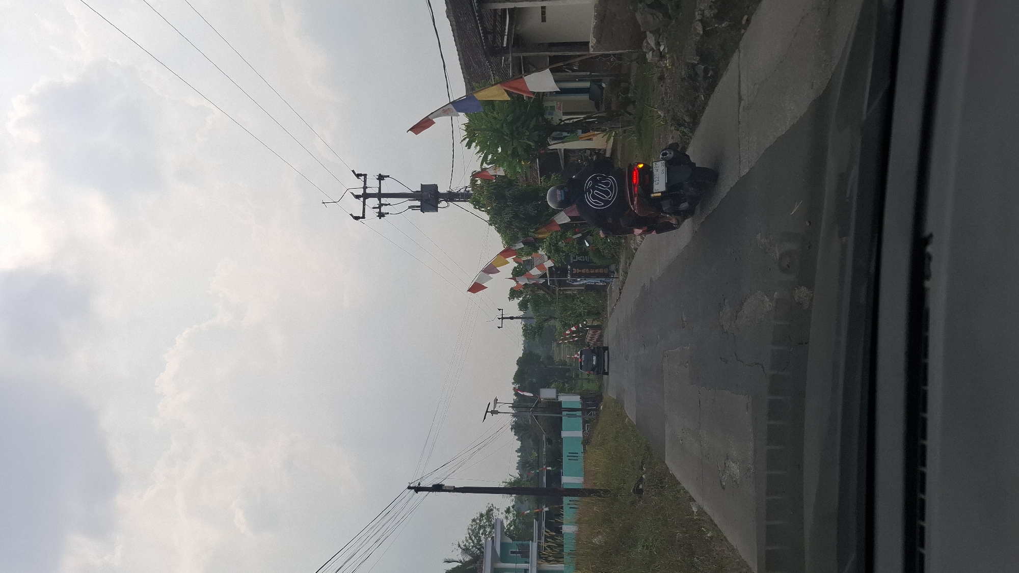 The image depicts a road in Cibinong Hilir, Indonesia, with Indonesian flags adorning the sides of the road. A black car is visible in the distance driving away from the camera, while a motorcycle with two occupants is closer to the foreground, also moving away. The sky is overcast with diffused clouds. Utility poles with wires are present along the road. The environment appears to be a semi-rural or suburban area with buildings and vegetation visible on both sides of the road. The road itself shows signs of wear and tear. The perspective is from inside a vehicle, looking forward through the windshield.
