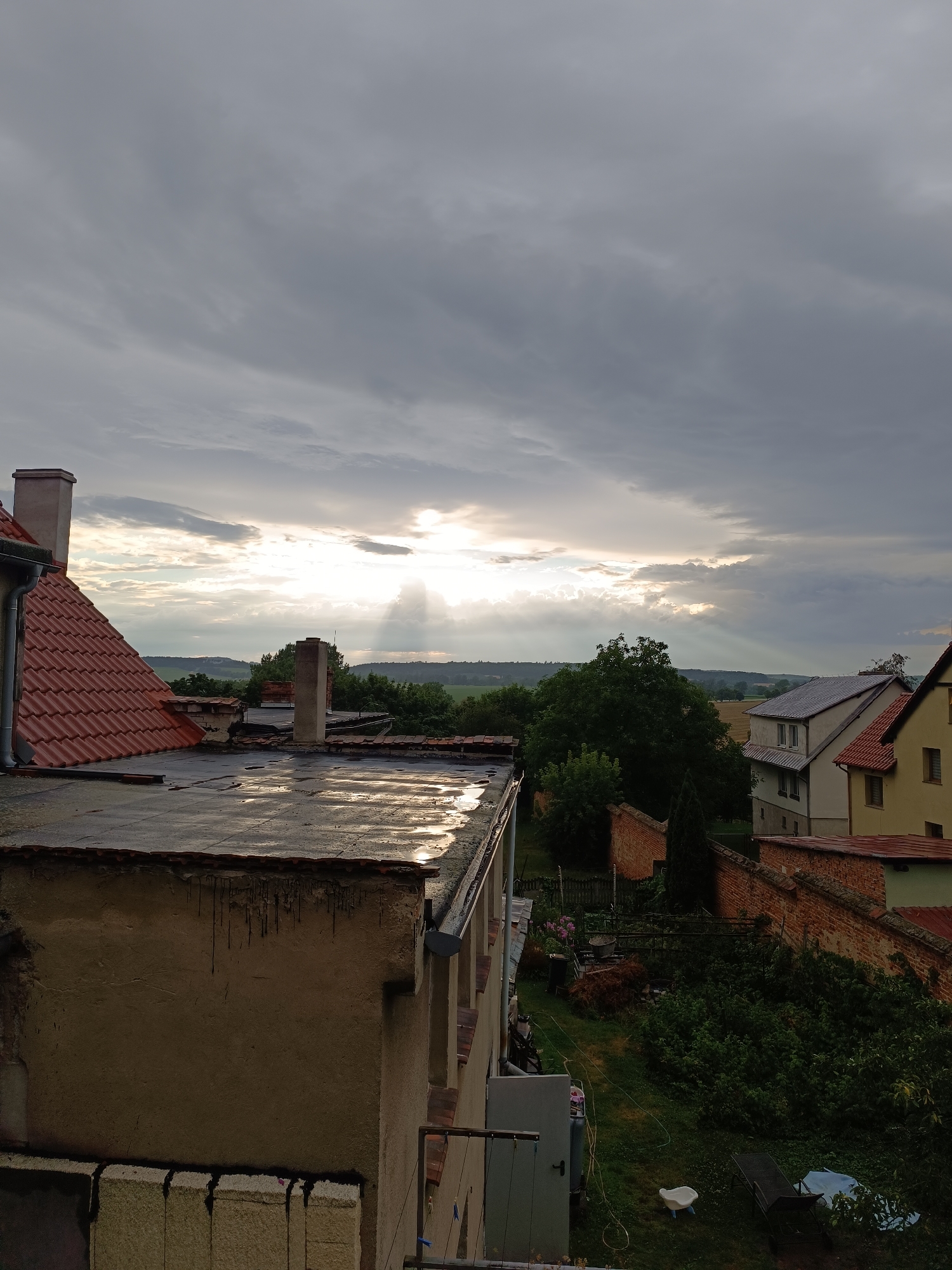 The image depicts a view from a rooftop in Niemcza, Poland, looking towards a rural landscape under a dramatic, cloudy sky. The foreground is dominated by the flat roof of a building with visible water pooling and reflecting the light. To the left, a portion of a steeply pitched red-tiled roof with a chimney is visible. Several other chimneys poke up from various buildings in the middle ground.
Beyond the immediate buildings, a lush green treeline stretches across the horizon, with rolling hills visible in the distance. The sky is filled with heavy, gray clouds, but shafts of sunlight are breaking through in the center, creating a dramatic light effect. On the right side of the image, residential buildings with tiled roofs and brick walls are visible, suggesting a residential area. A small garden with dense greenery and a brick wall is also present. There are no people visible in the image. The overall atmosphere is one of natural beauty contrasted with aging urban structures.
