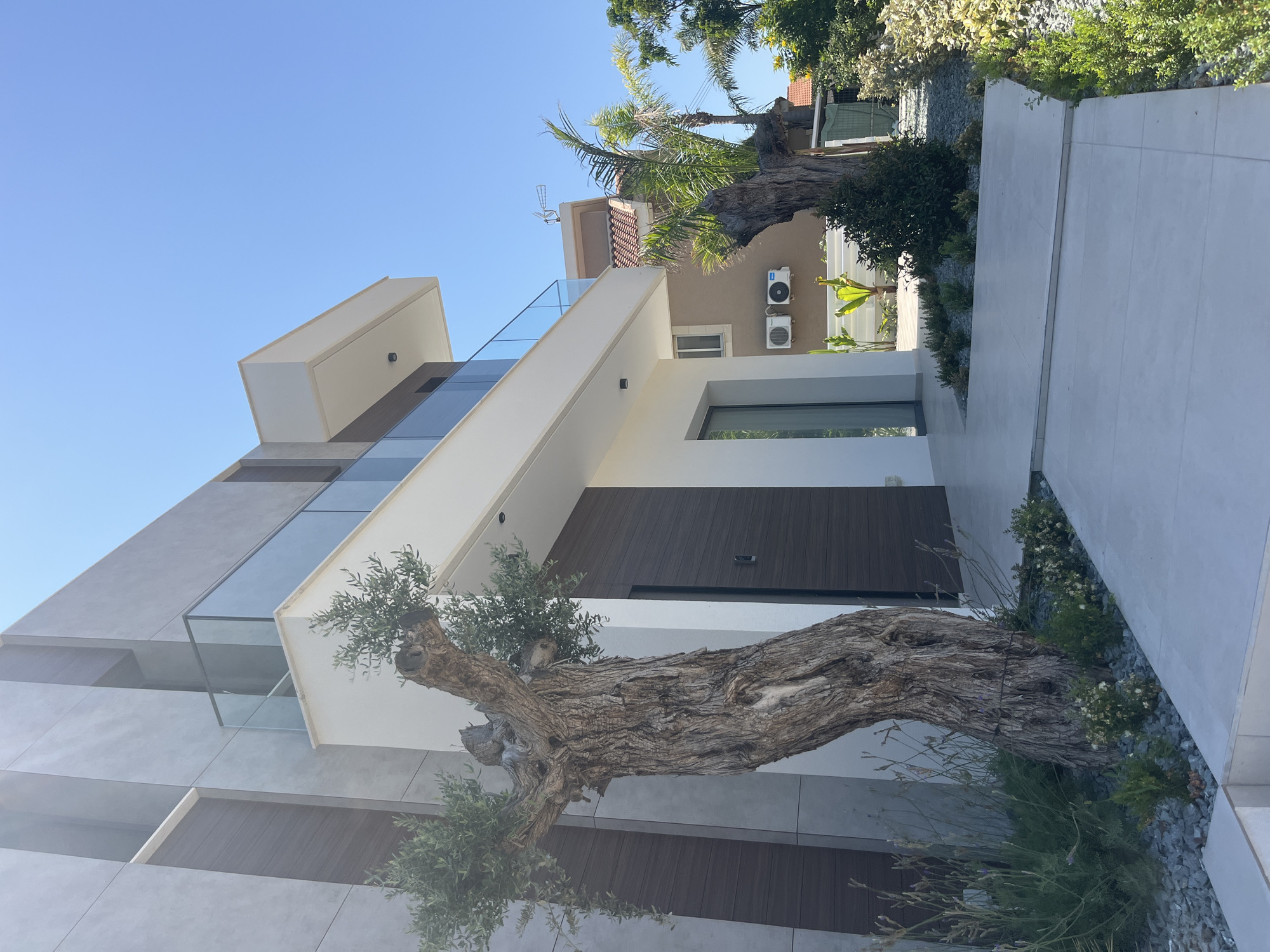 A modern house with a striking architectural design is shown in an exterior shot. The house features clean lines, a mix of concrete and wood-paneled facades, and large glass windows and balconies. A prominent, gnarled olive tree trunk extends horizontally across the foreground, its rough bark and sparse foliage creating a dramatic contrast with the sleek building. The setting is a residential area, with glimpses of neighboring houses and lush greenery. The sky is clear and blue, indicating a sunny day. Two outdoor air conditioning units are visible mounted on the beige wall of the house. The scene appears to be set in Moni Community, Cyprus. There is no visible text in the image.
