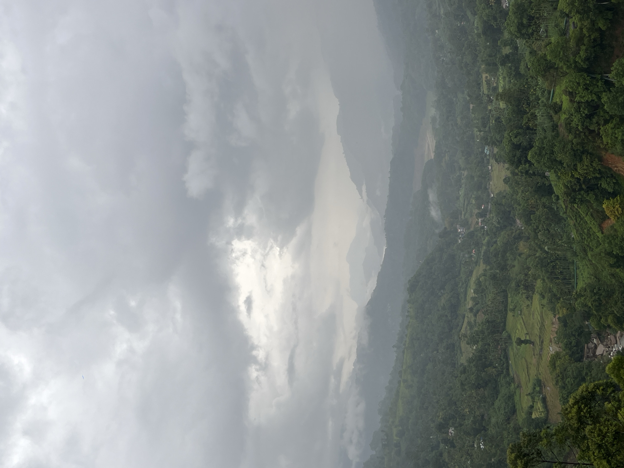 The image depicts a hilly landscape in Ramboda, Sri Lanka, under overcast skies. Dense vegetation covers the hillsides, with hints of rice paddies and small settlements scattered throughout. Mountains fade into the distance, obscured by mist and low clouds. The sky is dominated by gray clouds, with a patch of brighter light breaking through.