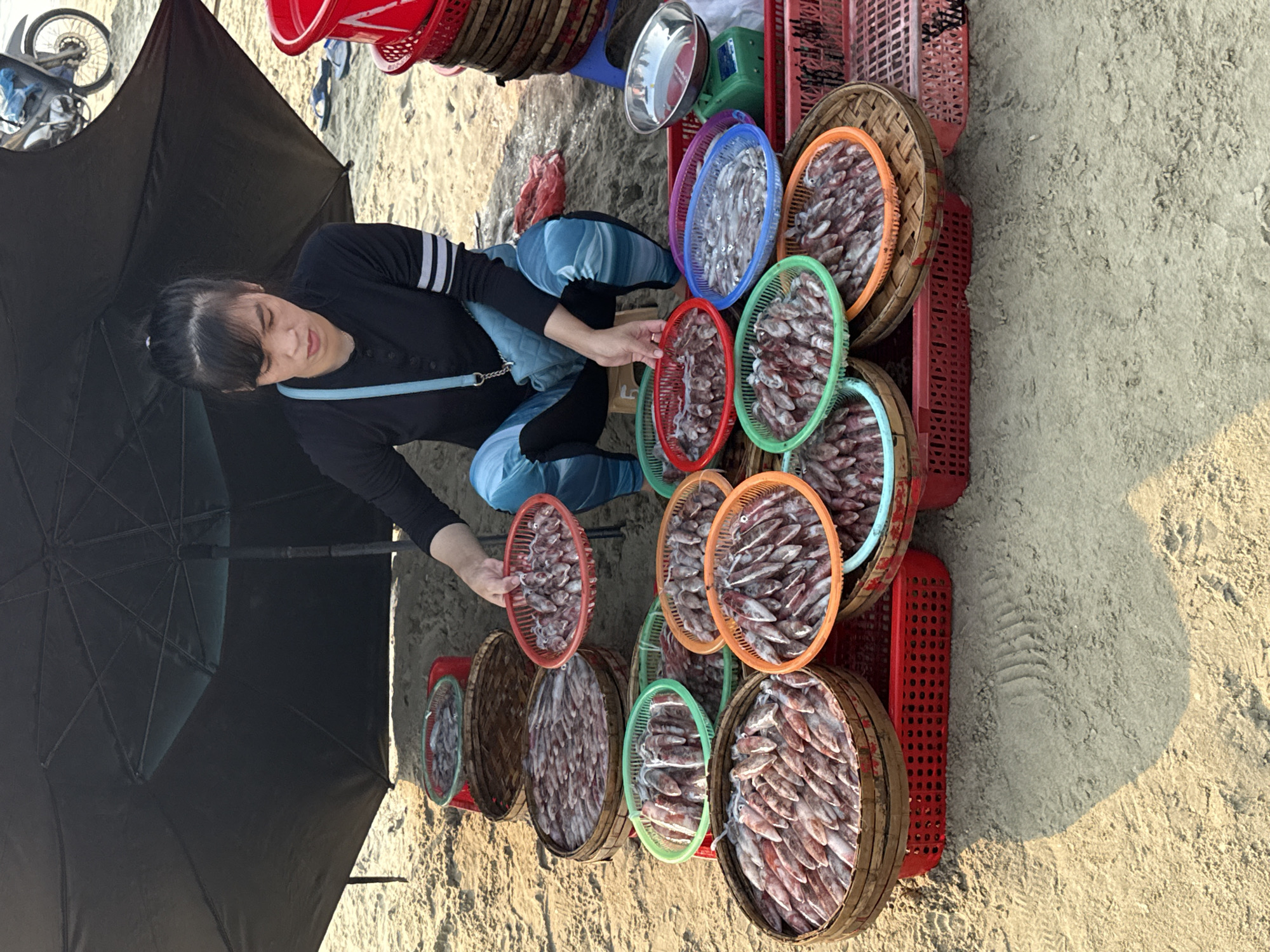 A woman is selling dried squid in Đà Nẵng, Vietnam. She is crouched under a black umbrella on a sandy surface, possibly a beach. The woman is wearing a black long-sleeved shirt with white stripes on the sleeves, light blue jeans, and a light blue shoulder bag. She is holding a basket of dried squid, which is among several other baskets filled with the same product. The baskets are placed on a red plastic crate. Additional objects include red plastic containers and a weighing scale. A motorcycle is partially visible in the background.