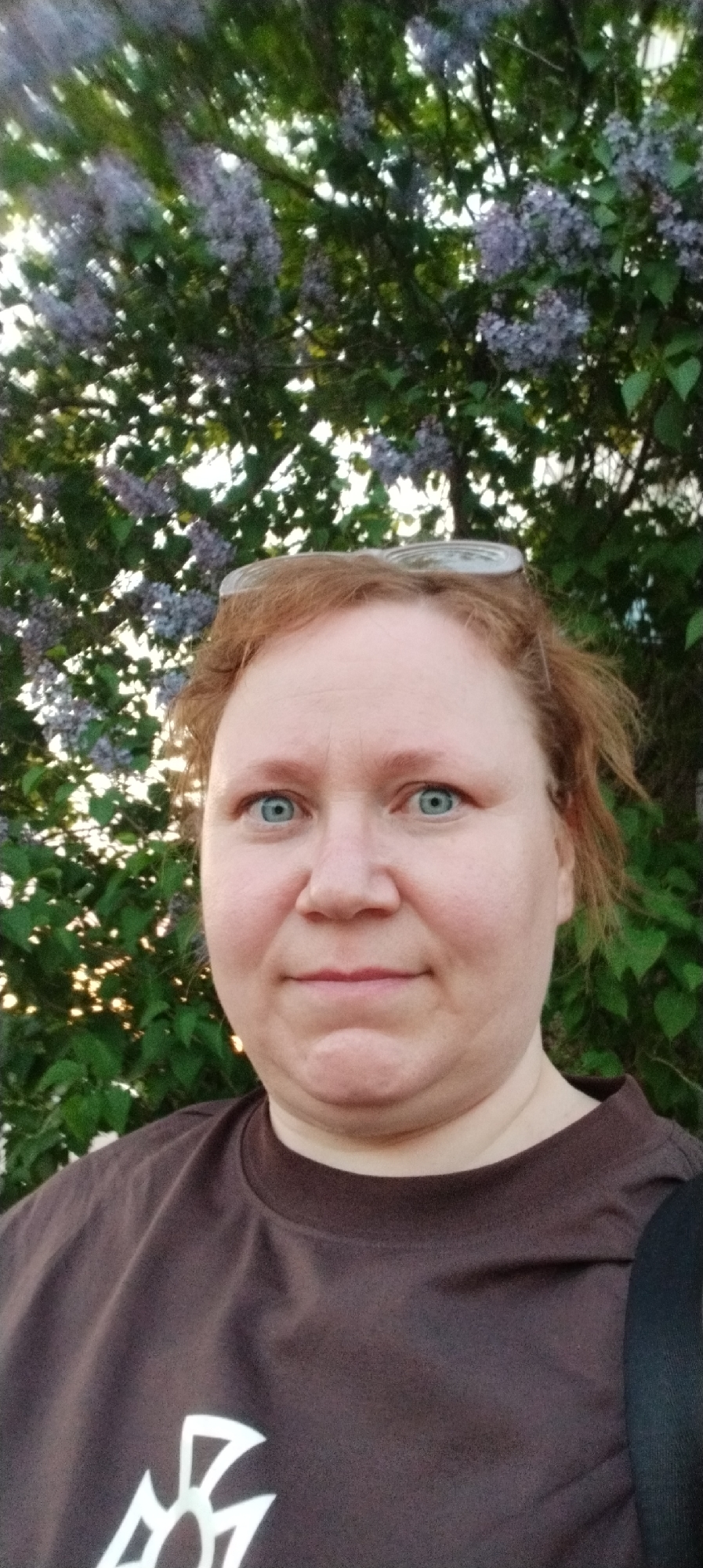 A woman with fair skin and blue eyes is photographed in front of a lilac bush. She has reddish-blonde hair and is wearing glasses perched on her head. She is wearing a brown t-shirt with a white design on the front. The background includes green foliage and blooming lilac flowers. The location is Chaikovsky, Russia.