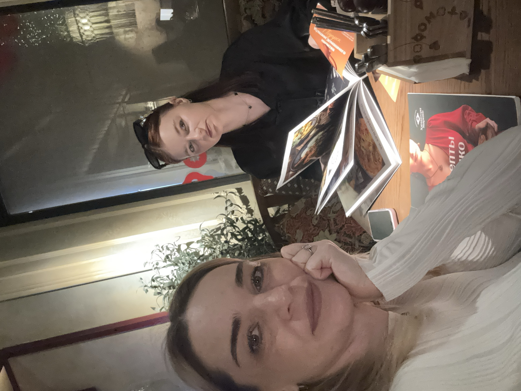 Two women are at a table in what appears to be a restaurant in Saint Petersburg, Russia, as indicated by the Cyrillic script on objects in the scene. The woman in the foreground is taking a selfie with her hand on her face. She is wearing a white top. The other woman, in the background, is dressed in black and is looking at a menu. Open menus are present on the table, as well as a napkin holder and what seems to be a book. The environment includes indoor plants.