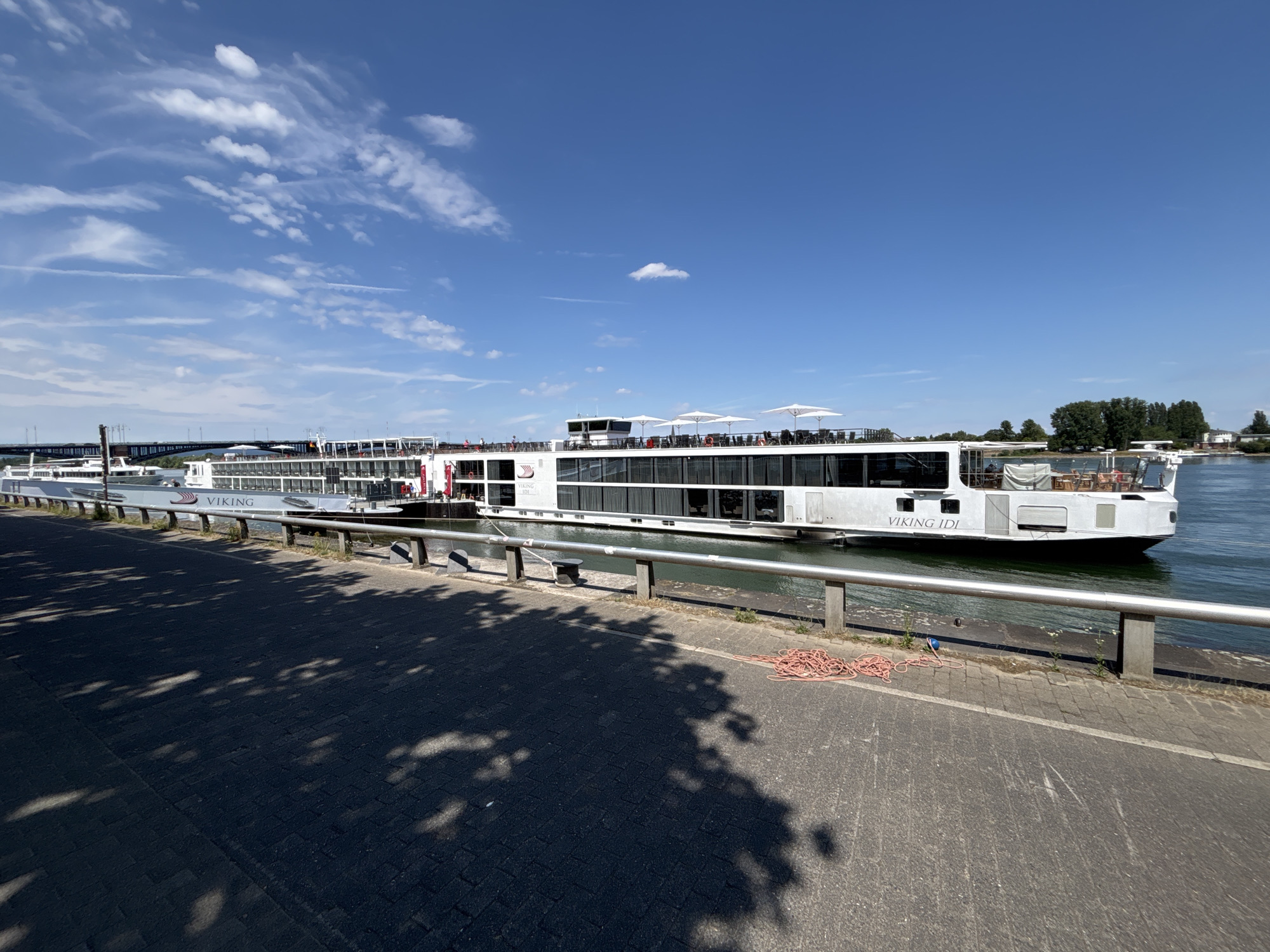 A long, white river cruise ship, named "VIKING IDI," is docked on a calm waterway under a bright blue sky with scattered white clouds. The ship features multiple decks with rows of windows and an upper deck with white umbrellas and outdoor seating. The port side of the ship is visible, with the name "VIKING" prominently displayed in large letters.
The foreground shows a paved walkway adjacent to a metal railing that separates the walkway from the water. Tree shadows are cast across the walkway, suggesting it is midday. On the walkway, a coiled orange rope lies near the railing. In the distance, a bridge and trees can be seen on the far bank of the river. The scene appears to be a sunny day in a European city, likely Mainz, Germany, given the context. There are no people clearly visible on the ship or the walkway, contributing to a sense of peaceful stillness. The overall impression is one of a serene, sunny day at a riverside.