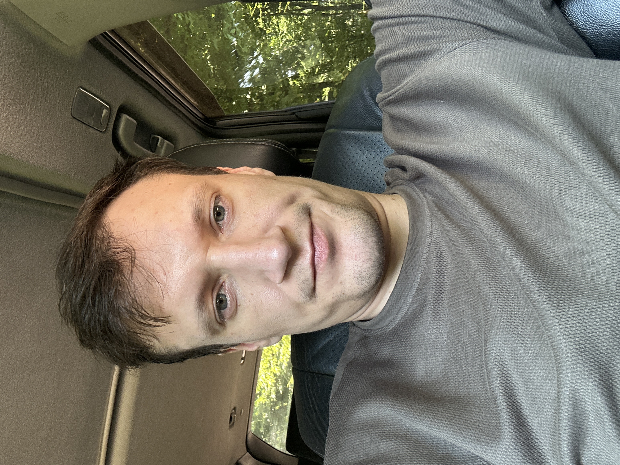 A man is captured in a selfie taken inside a vehicle. He has short, dark brown hair, light skin, and blue eyes. He is wearing a dark gray, long-sleeved shirt. The man is looking directly at the camera with a neutral expression. The background shows the interior of the car, including a black leather seat and a window that reveals green trees and foliage, suggesting an outdoor, possibly rural setting. The image does not contain any information or context that would indicate a location in the city of Shatava, Ukraine.