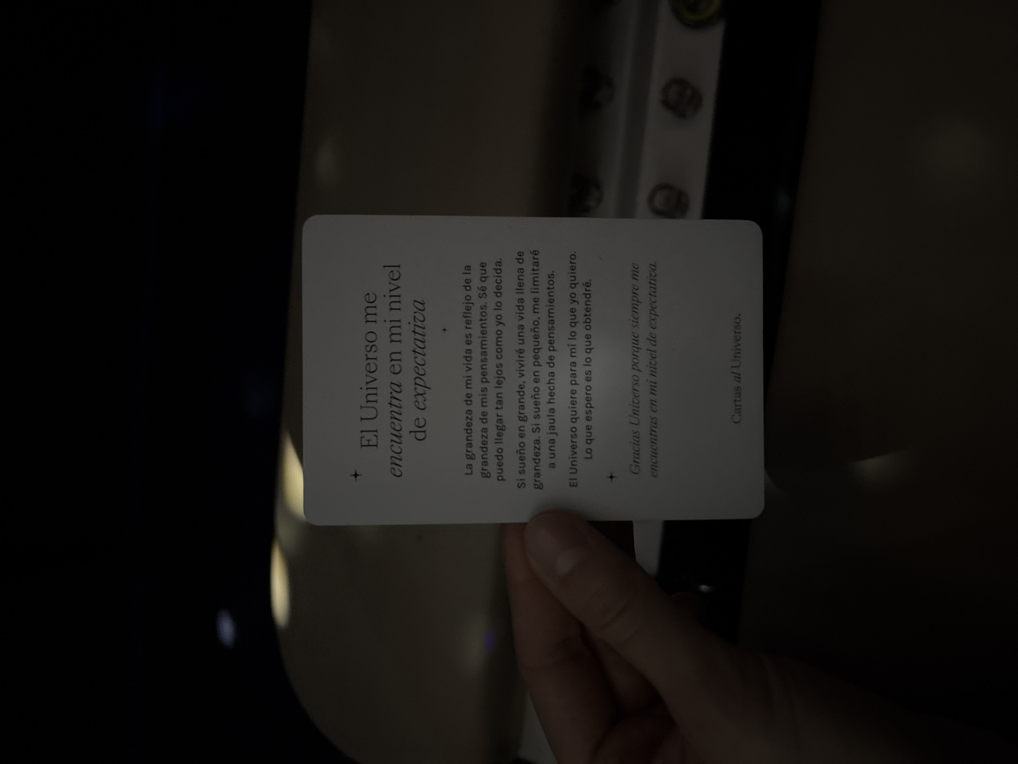 A hand is holding a white card with Spanish text on it. The card has a dark background and is partially obscured by the hand. The text on the card reads:
"El Universo me
encuentra en mi nivel
de expectativa
La grandeza de mi vida es reflejo de la
grandeza de mis pensamientos. Sé que
puedo llegar tan lejos como yo lo decida.
Si sueño en grande, viviré una vida llena de
grandeza. Si sueño en pequeño, me limitaré
a una jaula hecha de pensamientos.
El Universo quiere para mí lo que yo quiero.
Lo que espero es lo que obtendré.
Gracias Universo porque siempre me
encuentras en mi nivel de expectativa.
Cartas al Universo."
The environment is dark, with some light reflecting off the card and the hand. The background appears to be a blurred interior space, possibly a vehicle or a room. There are no discernible landmarks or contextual clues that would place this image in Mexico City, Mexico.