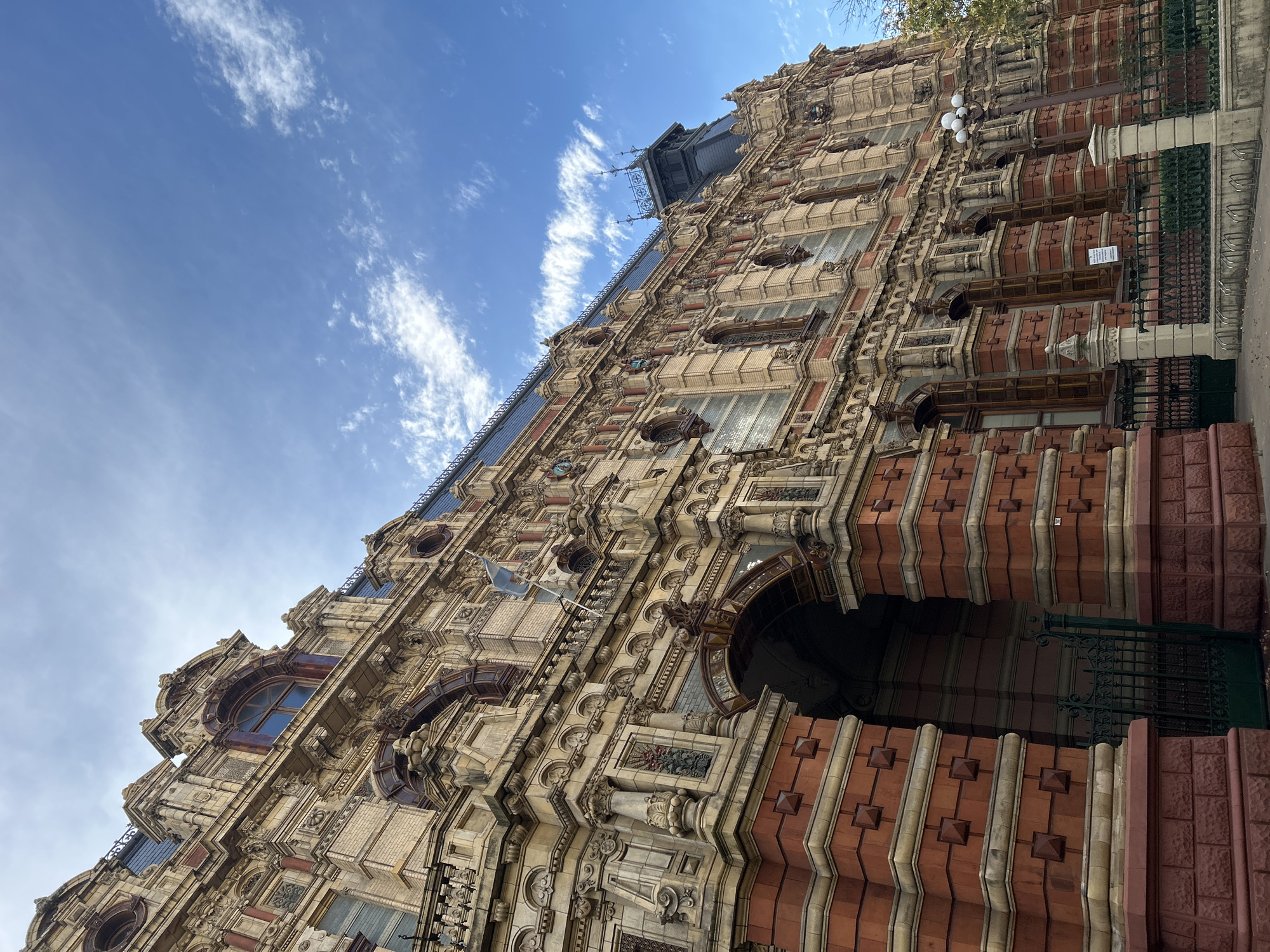A low-angle shot of the Palacio de Aguas Corrientes in Buenos Aires, Argentina. The building, shot from the ground, showcases its ornate facade, detailed brickwork, and numerous windows. The architectural style is elaborate, with protruding elements and decorative features. The sky is visible in the background, with blue patches and scattered clouds. A portion of the sidewalk and a wrought-iron fence appear in the foreground. No people are visible.