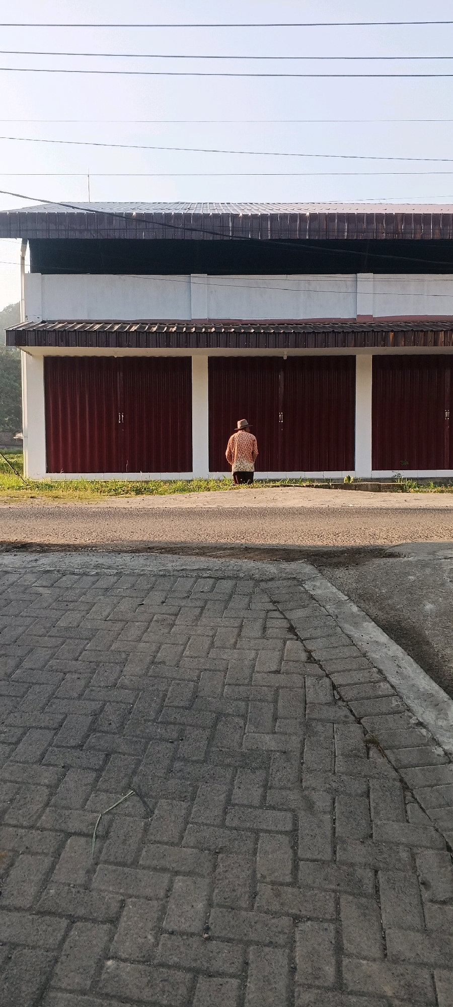 A person wearing a hat and a patterned shirt kneels on the ground in front of a building with several red rolling doors. The person's back is to the camera. The building appears to be a commercial structure with a white facade above the doors and a dark-colored roof. A paved area with interlocking bricks is in the foreground, separated from the building by a strip of asphalt and a small grassy area. The scene is illuminated by natural light, suggesting it is daytime. The background shows a slightly hilly landscape and a clear sky with overhead wires.