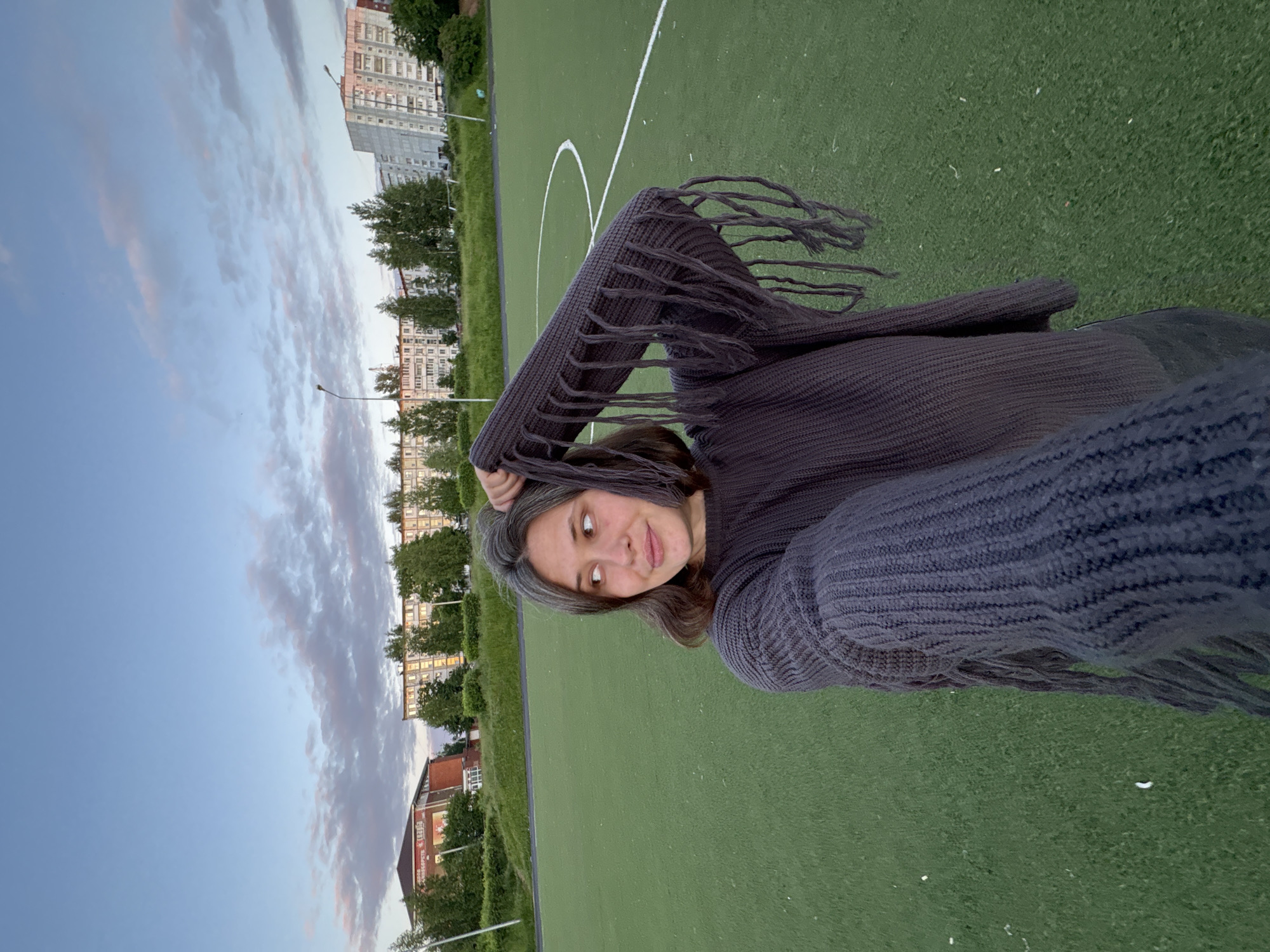 A female individual, likely in her 20s or 30s, with shoulder-length dark brown hair displaying gray strands, is depicted in a selfie taken in Chaikovsky, Russia. She wears a dark gray, long-sleeved knitted sweater featuring distinct fringe elements along the sleeves. Her right arm is raised, with her hand positioned near the back of her head and hair. She looks towards her right (viewer's left) with a slight smile. The subject stands on a green artificial turf sports field, characterized by a white curved line segment on the right side. In the background, several multi-story residential buildings are visible, including one red-brick structure on the far left and lighter-colored apartment blocks, some with illuminated windows. Numerous green trees and bushes are situated between the field and the buildings. The sky is partly cloudy with a clear blue base and scattered, wispy clouds exhibiting pink and purple hues, indicating dusk or early evening. Two streetlights are also present in the background.