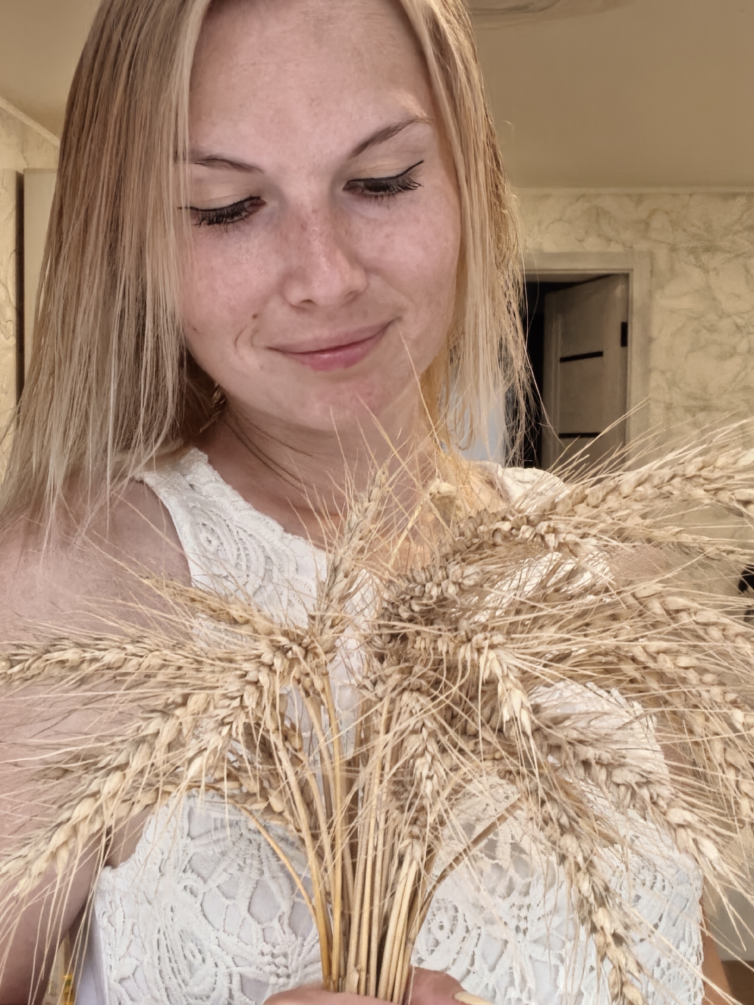 A medium shot photograph shows a blonde woman with light skin and freckles holding a bouquet of wheat stalks. She is wearing a white lace top and looking down and to her right with a slight smile. The background is blurry and appears to be an indoor setting with a marbled wall and a doorway. The lighting is soft, casting a warm glow on the subject. The image was taken in Sterlitamak, Russia.