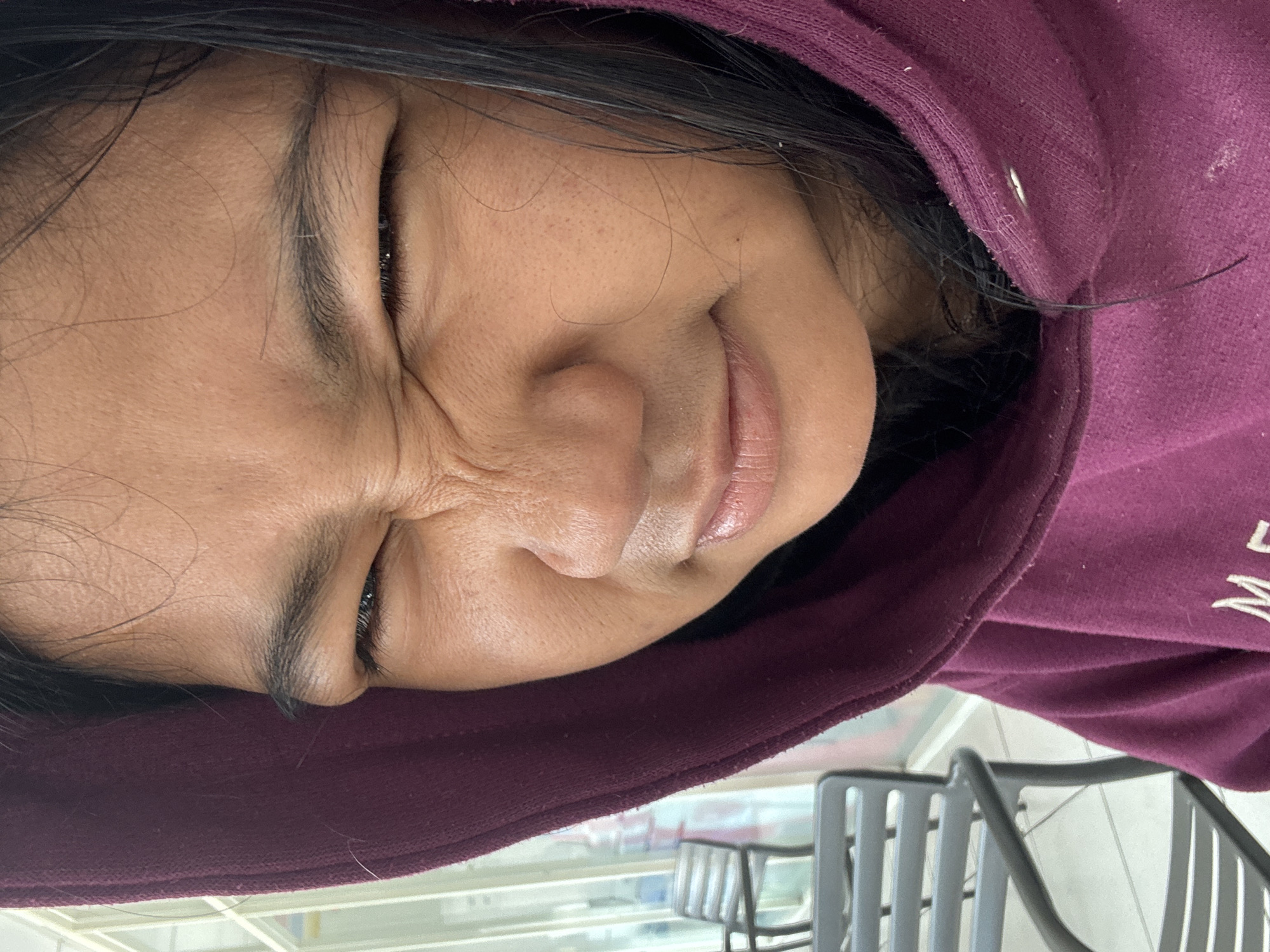 A close-up, slightly angled shot shows the face of a young person wearing a dark purple hooded sweatshirt. The person's face is scrunched up, with eyes squinted and eyebrows furrowed, suggesting a moment of discomfort or displeasure. Their mouth is closed with a slight pout. Dark hair frames their face. The background is blurred but suggests an indoor setting with outdoor elements visible through a window or glass partition, including outdoor seating with grey chairs. The overall impression is informal, possibly taken in a cafe or a public seating area in North Jakarta, Indonesia.