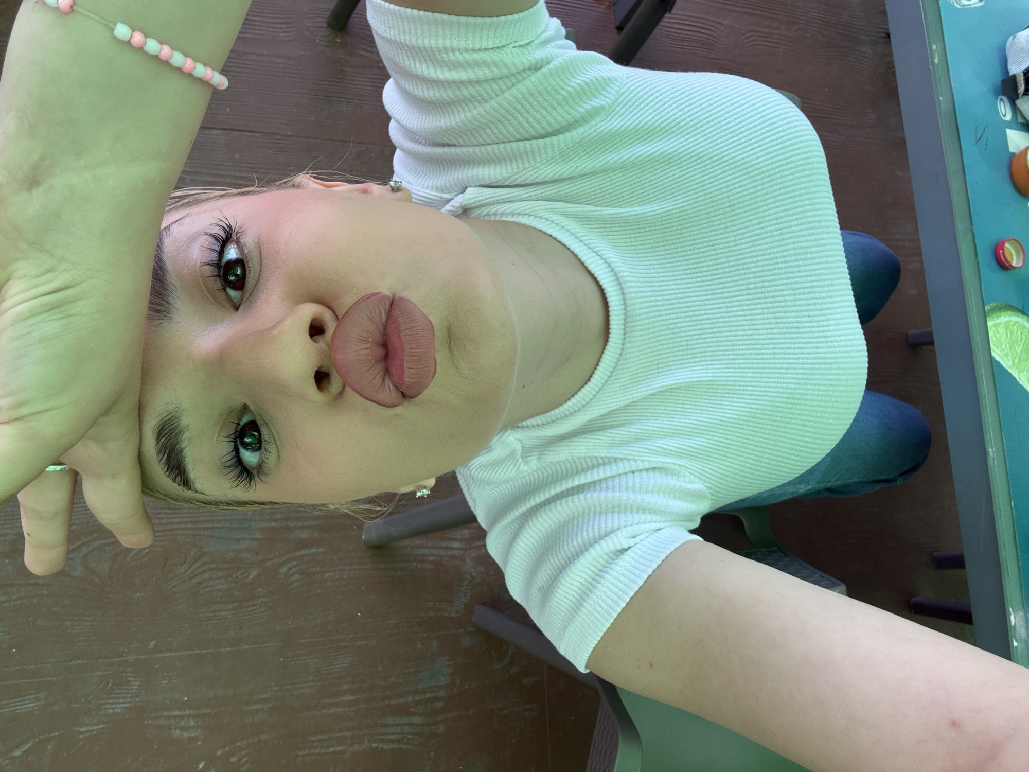 A high-angle selfie captures a young individual, likely female, striking a "duck face" pose with pursed lips and looking directly at the camera. The person has fair skin, prominent dark eyelashes, and neatly groomed eyebrows, with a subtle pink flush on the cheeks. They are wearing a white, ribbed, long-sleeved top and blue jeans, with a delicate pink and white beaded bracelet on one wrist and a small stud earring visible.
The setting appears to be outdoors, possibly on a patio or deck, characterized by a brown wooden floor or decking. Parts of dark metal chair legs are visible. To the right, a light blue-green table is present, holding several items including a red bottle cap, an orange spherical object, and a slice of lime or similar fruit. The scene is brightly lit by natural daylight. No discernible text is visible in the image.