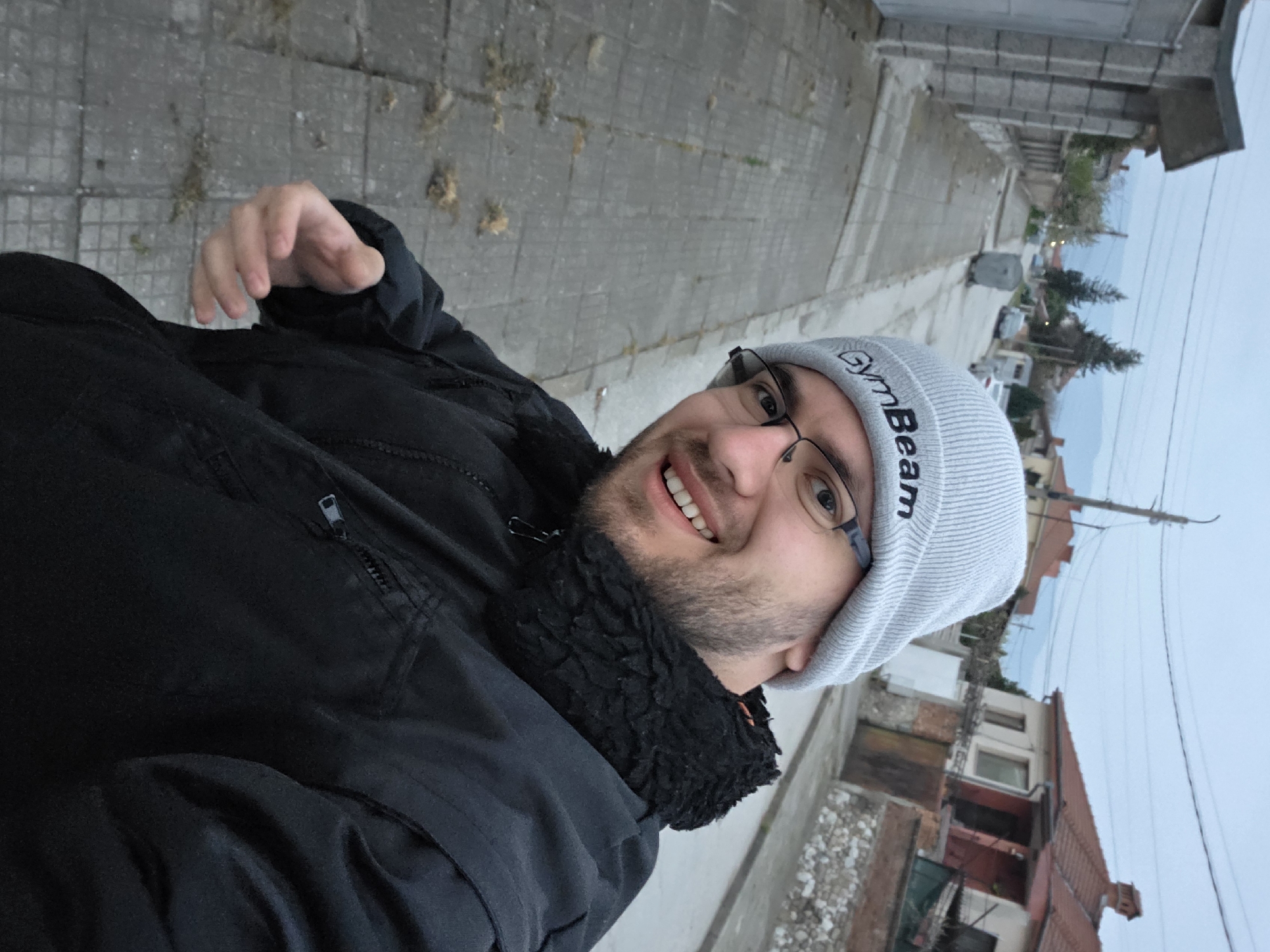 A smiling man wearing glasses and a gray beanie with the word "GymBeam" stands outside, facing the camera. He is wearing a black jacket with a fur-lined collar and a zipper on the left side. The background shows a residential area with concrete sidewalks and a row of houses, including some with red tile roofs. The sky is overcast. The picture appears to be taken in Yagodovo, Bulgaria.