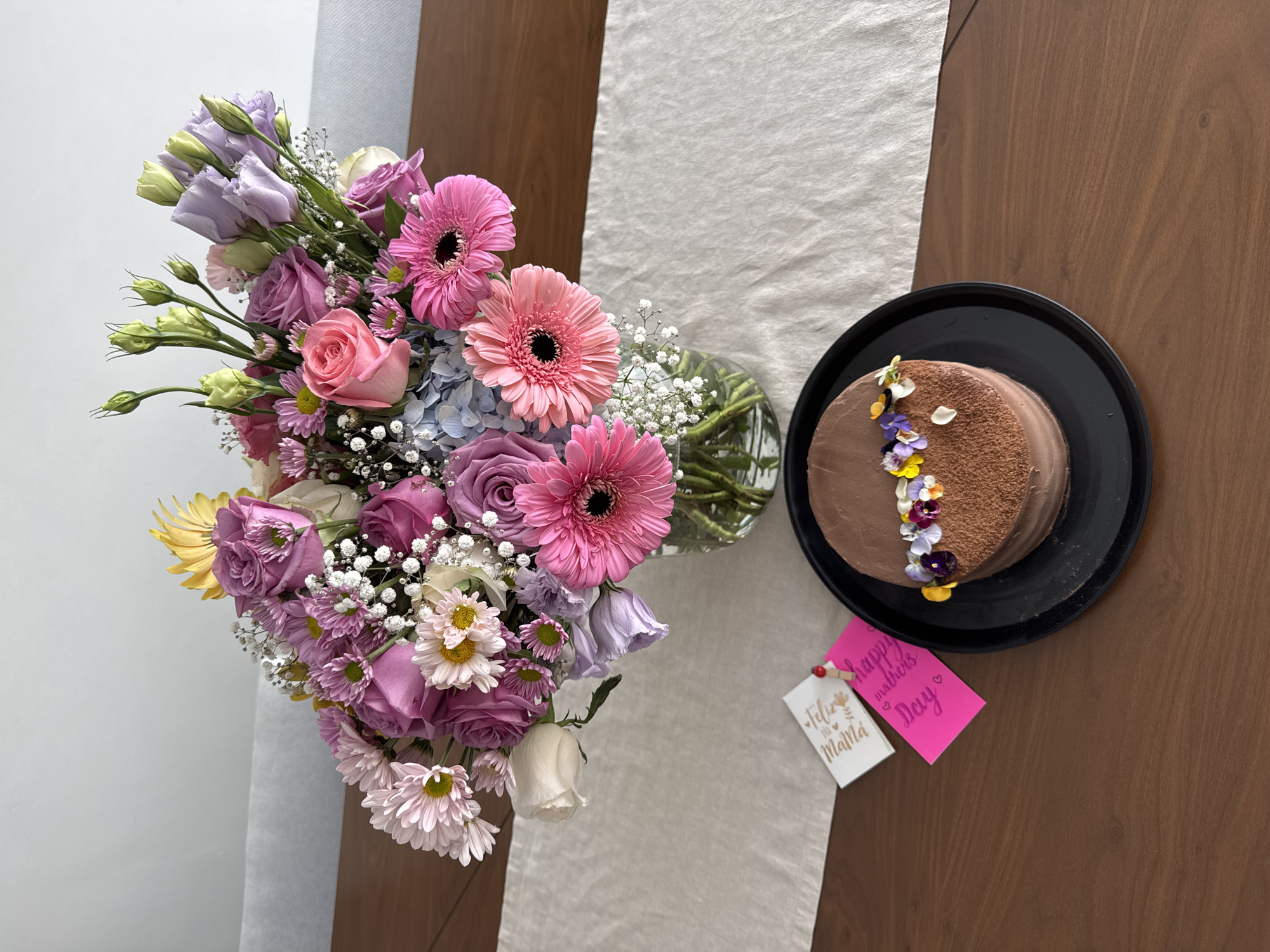 A top-down shot shows a table with a vase of flowers and a cake. The flowers are a mix of pink gerberas, purple and pink roses, white daisies, and baby's breath. They're arranged in a clear glass vase. A chocolate cake sits on a black plate. The cake is decorated with a line of edible flowers. Two small cards lay next to the cake. One says "Feliz Dia Mama" and the other says "Happy Mother's Day". The table has a wooden surface, covered by a white table runner. The setting suggests a celebration, most likely Mother's Day. The location is Puebla, Mexico, based on the Spanish text on the card.