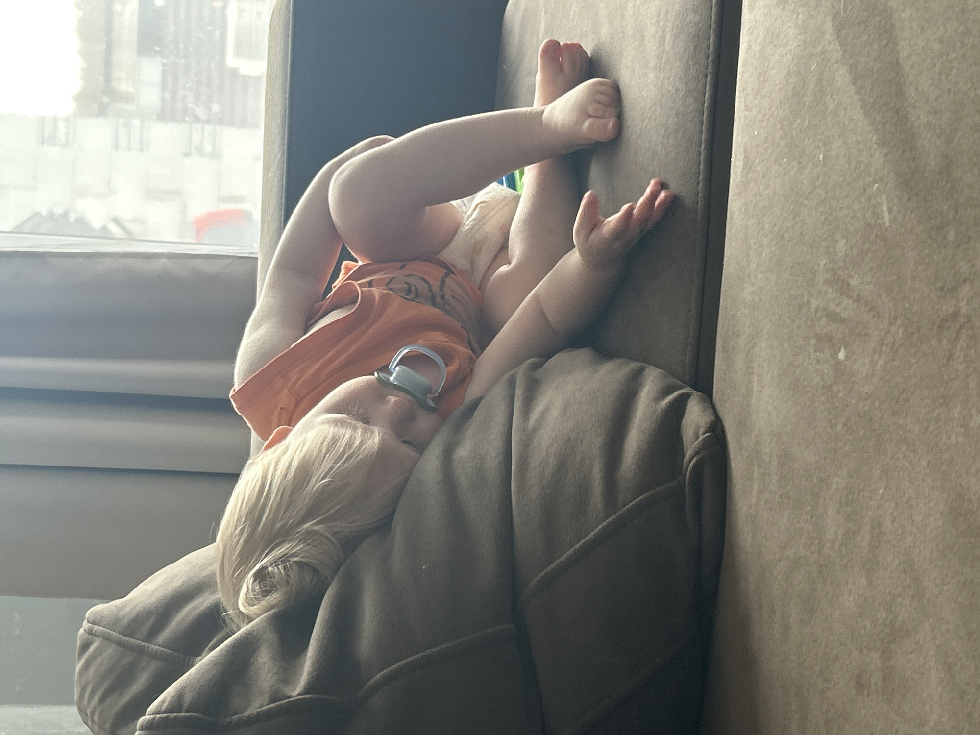A child is asleep on a brown couch with their legs up the side of the couch. The child has blonde hair and is wearing a pacifier. The background shows a window overlooking a city, possibly Chaikovsky, Russia. The child is wearing a diaper and an orange t-shirt. The child is lying on a couch cushion.