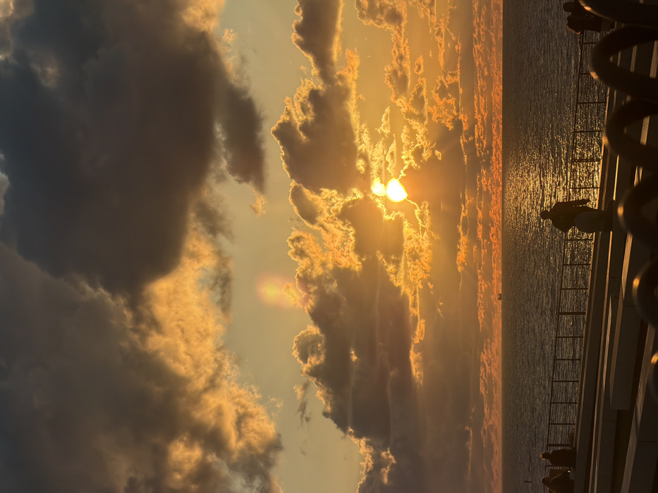 A vibrant sunset casts golden hues across a sky filled with dramatic clouds, some dark and looming, others radiant with light. The sun, partially obscured by clouds, shines brightly, creating beams of light that illuminate the water below. A railing runs along the right side of the frame, with a person leaning on it, silhouetted against the glittering sea.