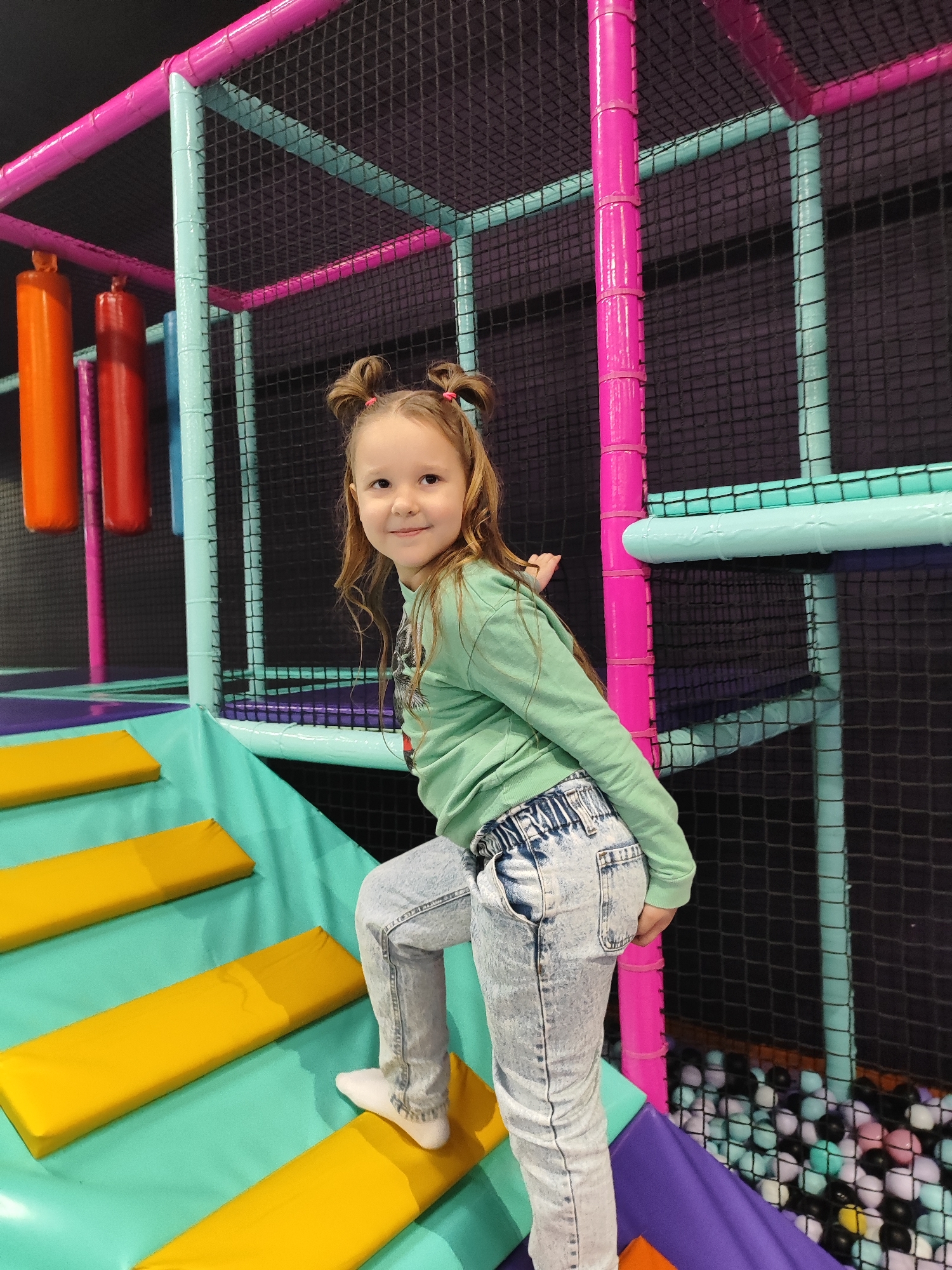 A young girl with brown hair styled in two small buns with pink hair ties is climbing yellow stairs in an indoor playground. She is wearing a light green long-sleeved shirt, light blue denim jeans, and white socks. The playground features colorful padded structures in pink, blue, and purple, with a black netting enclosure. There are orange and red padded cylinders hanging in the background. A ball pit with various colored balls is visible near the stairs. The location is likely a children's entertainment center in Krasnodar, Russia.