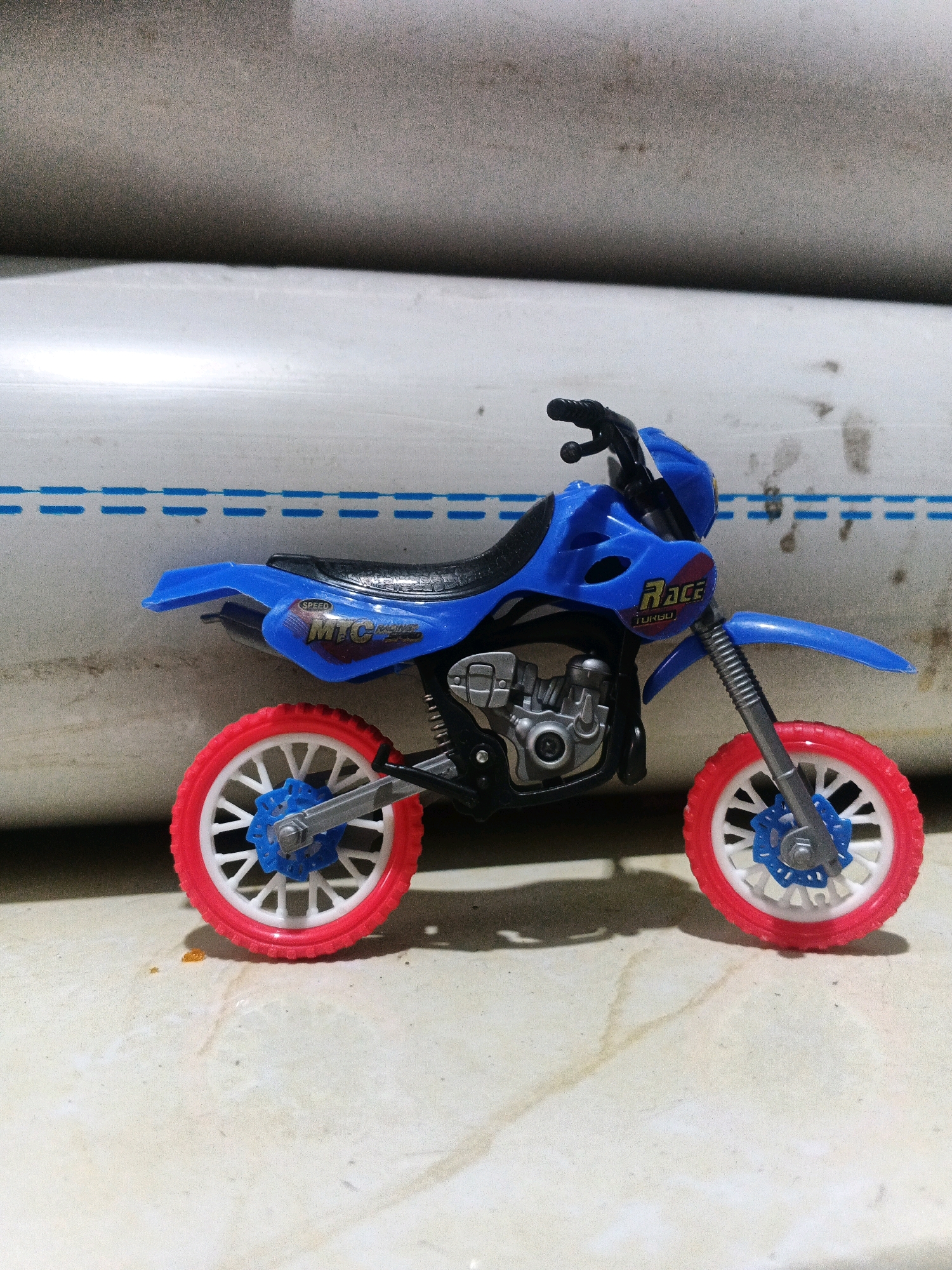 A blue toy dirt bike is positioned on a beige, textured surface. The bike features red tires with white spokes and blue hubs. The front fender is blue, and the handlebars are black. The side of the bike has decals that read "SPEED" and "MTC RACING TURBO." In the background, a large white cylindrical object with blue dashed lines running horizontally is visible. The lighting suggests it is daytime. This scene is located in Kadu, Indonesia.