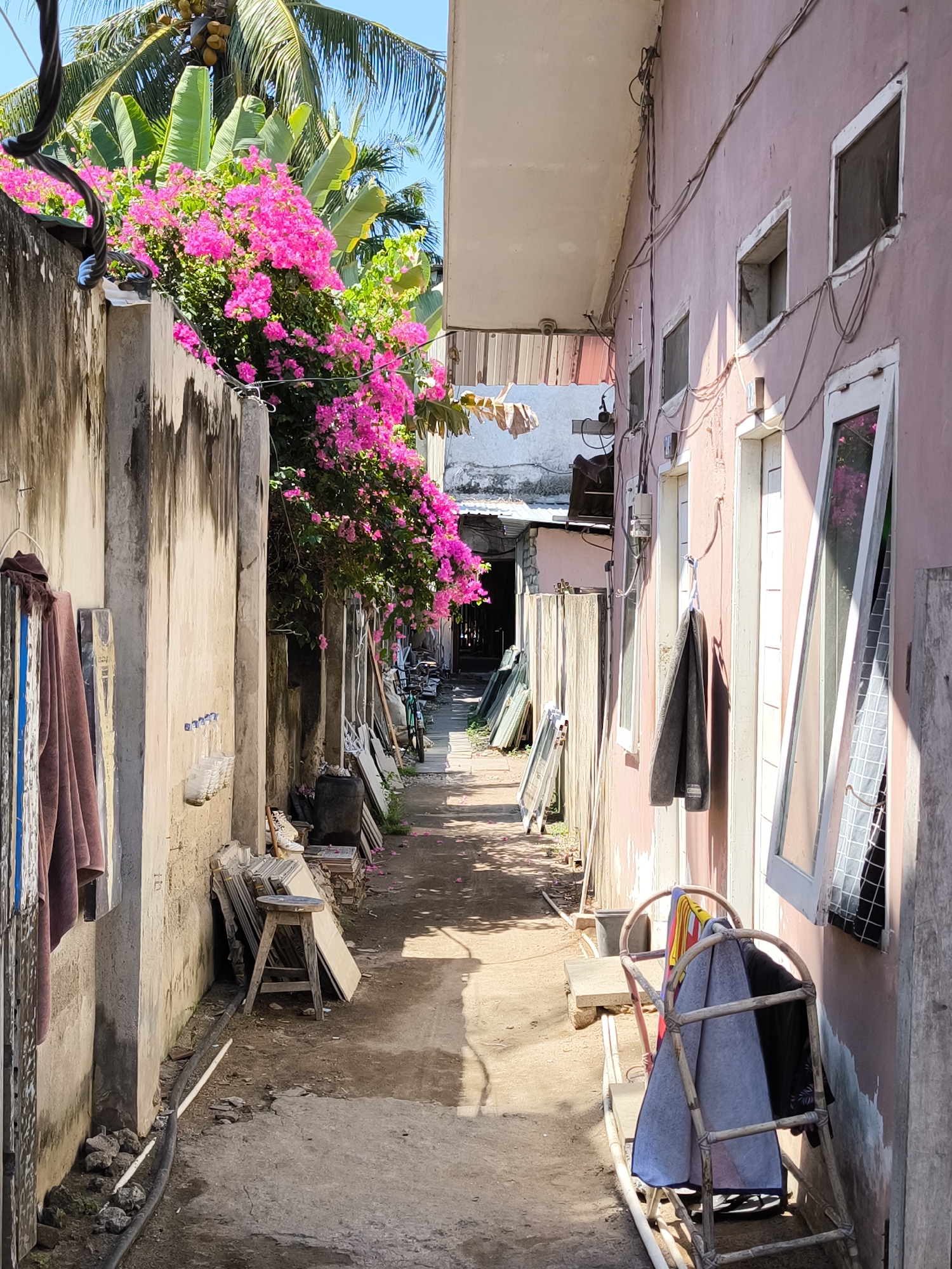 The image depicts a narrow alleyway in an urban environment in Unknown, Indonesia. The alley is flanked by a tall, weathered concrete wall on the left and a pink painted building on the right. Vibrant pink bougainvillea cascades over the wall on the left, partially obscuring the sky. The alley is littered with construction materials, including stacks of tiles, wooden planks, and pieces of glass. An old wooden stool sits near the left wall. Laundry, including towels and clothing, hangs from both walls. In the background, the alley leads to a shadowed area. A bicycle is visible in the distance. The ground is a mix of concrete and dirt. The overall impression is one of a lived-in, somewhat cluttered urban space.