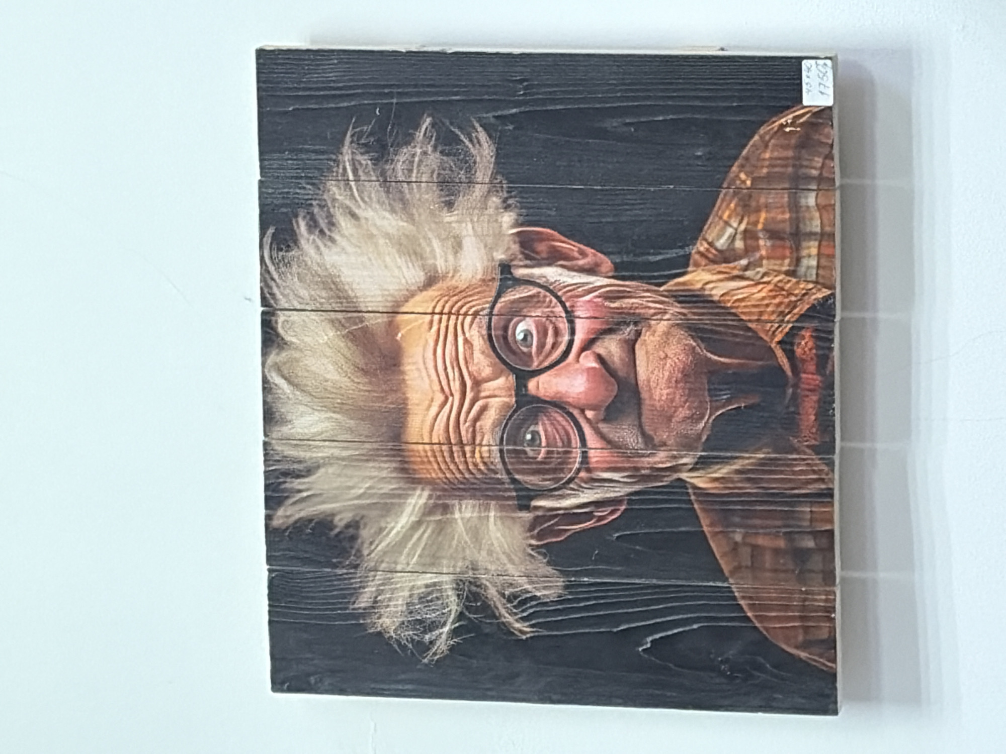 The image shows a rectangular wooden plank picture hanging on a white wall. The picture depicts an elderly man with white, fluffy hair, eyeglasses, and wrinkled skin. He is wearing a plaid shirt. The background is a dark, wood-grain texture. A small, white sticker with handwritten numbers is visible in the upper right corner of the picture. The image appears to be taken indoors. There is no indication in the image that would confirm its location in Izhevsk, Russia.