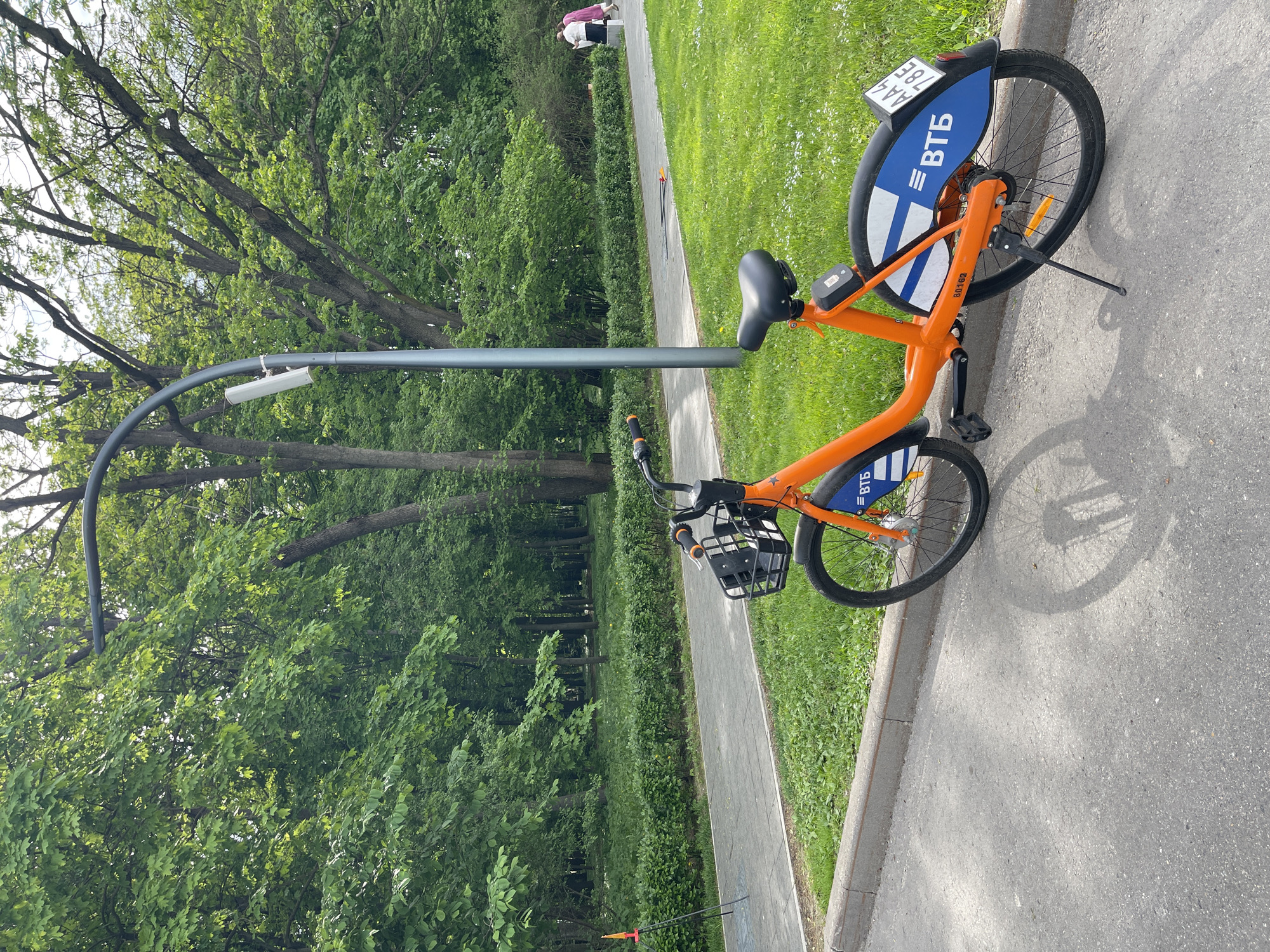 Here's a detailed description of the image:
The image shows an orange VTB bike standing on a paved path. The bike has a black basket in the front. The bike has a blue and white fender over the rear wheel, with a license plate above it. A gray metal structure that extends over the path is visible behind the bike. A sidewalk runs alongside the path. A grassy area with trees is visible to the left and in the background. Two people are standing on a path in the background.