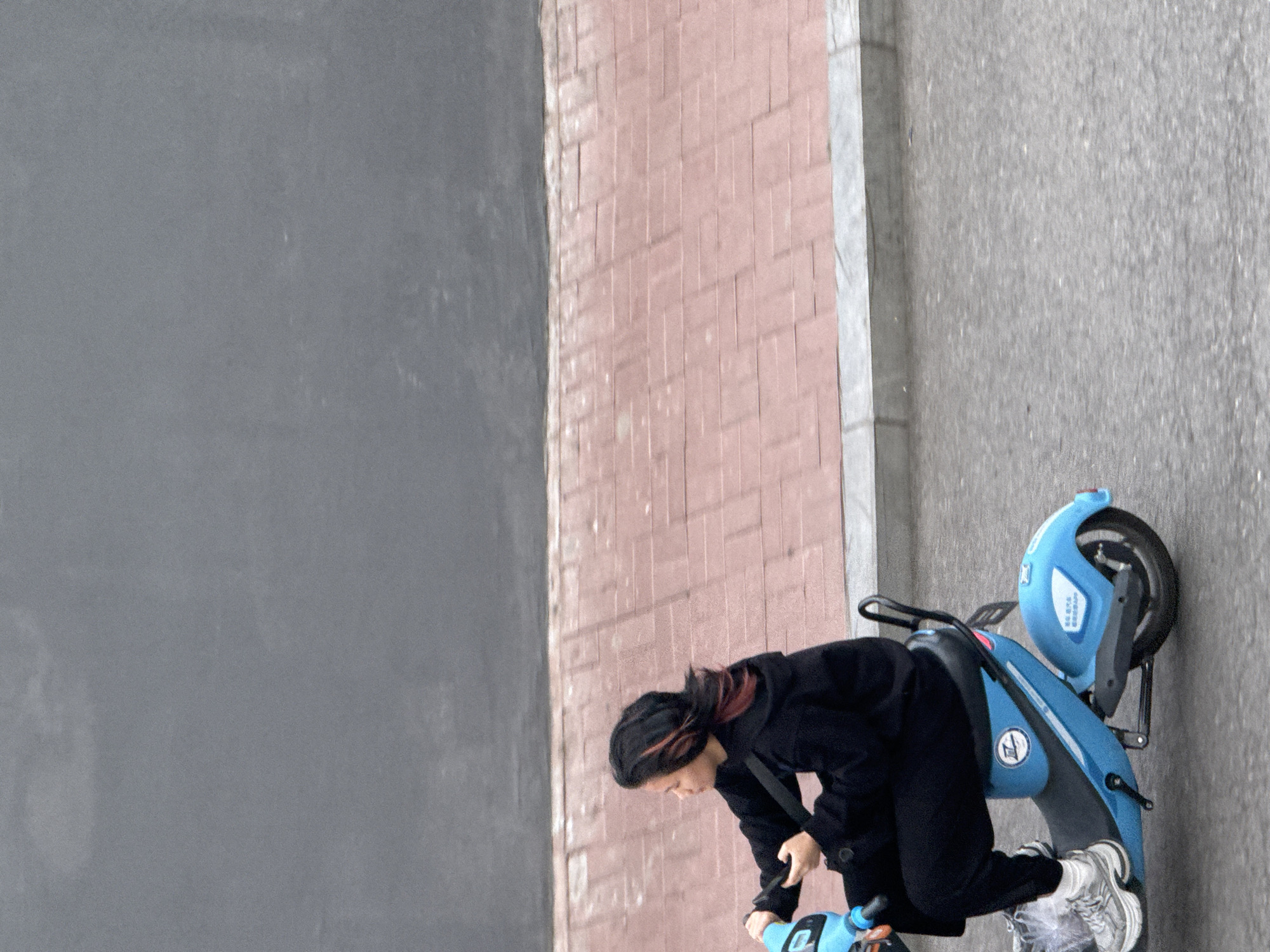 A person is bent over, interacting with a blue scooter parked on a sidewalk in Zhongtun, China. The sidewalk is bordered by a brick wall and a gray wall. The person is wearing a black jacket and pants, white socks, and gray sneakers. The blue scooter is partially visible, showing its design with a wheel and various parts. The scene appears to be outdoors, with natural lighting.