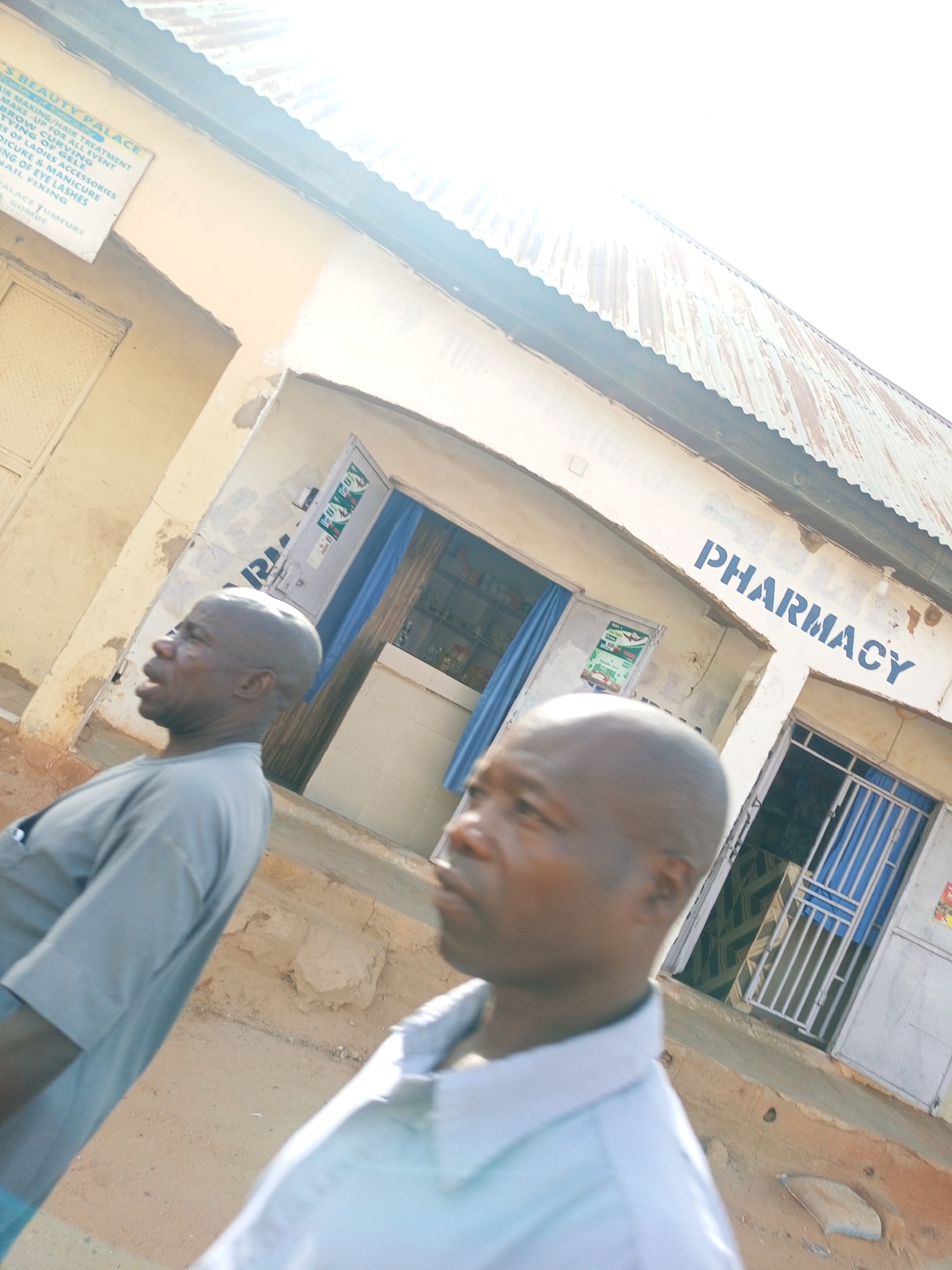 The image depicts two bald, dark-skinned men in the foreground, both wearing collared shirts and looking towards the left. Behind them, a building is visible with the word "PHARMACY" painted in blue on its facade. There are two doorways into the building, one with a blue curtain and another with a white barred gate. A sign on the left side of the image reads "S BEAUTY PALACE", located in "TUMFURE GOMBE". The building has a corrugated metal roof.