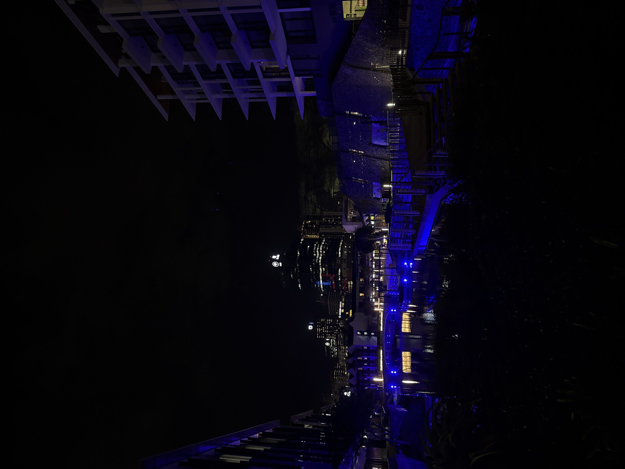 At night in Cape Town, South Africa, a high-angle view shows city buildings reflected in dark water. Above the water, blue lights line a bridge. Building architecture includes horizontal white planes at the top left. Below the central horizontal band is a blurred cityscape. The bottom third of the image shows building architecture with blue lights. The right side of the frame includes dark foliage.