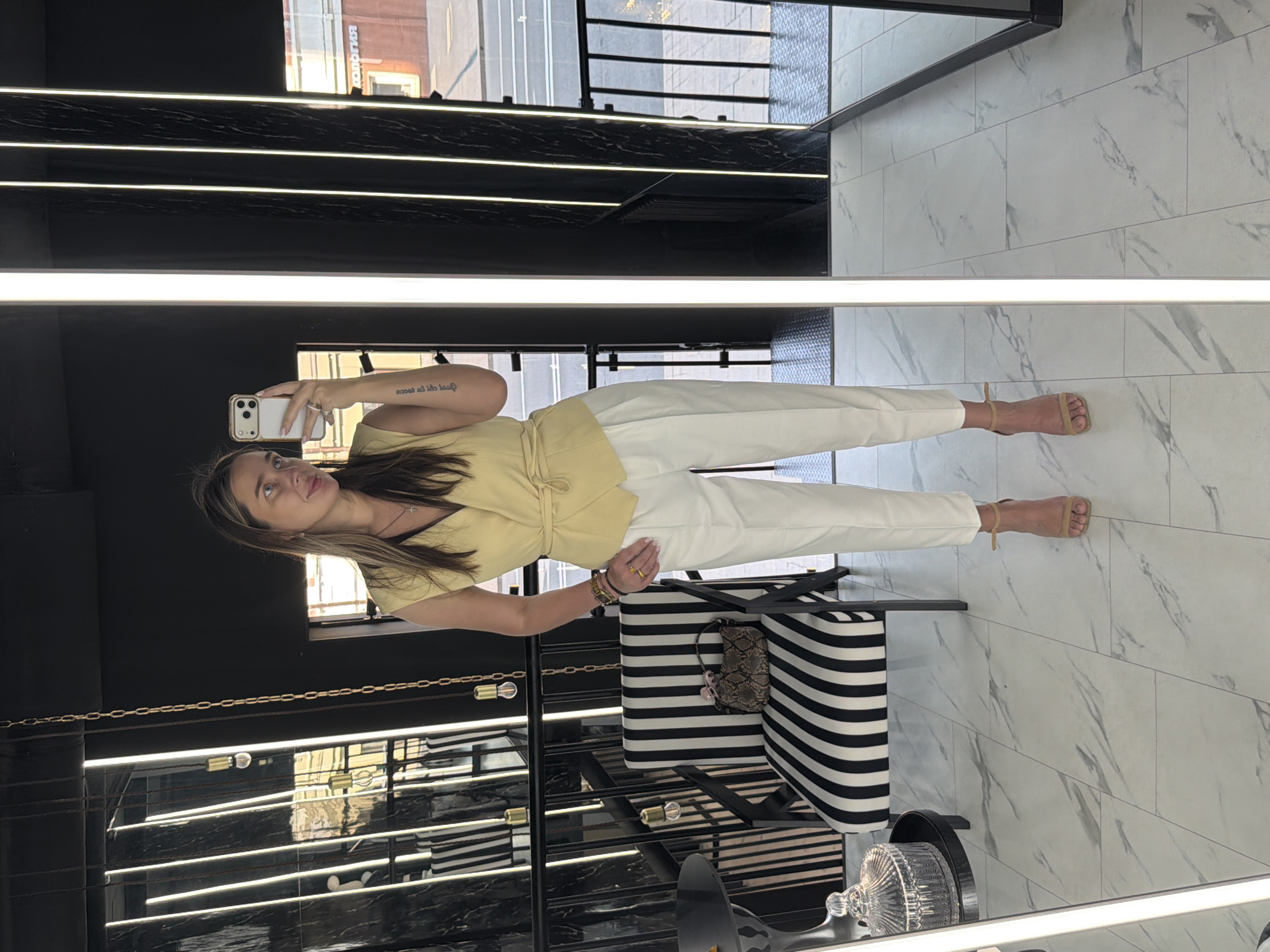The image shows a full-length view of a woman taking a selfie in a mirror, which is capturing a well-lit interior space. The woman is wearing a light yellow top and white pants with strappy sandals. She is holding a white smartphone in her left hand.
The interior features a black and white striped chair or bench, and a small purse is resting on it. The background suggests a modern setting with black walls and illuminated horizontal stripes. The floor is covered with white marble tiles. The location appears to be in a city, with an external view visible through a window or possibly reflected in another mirror above.