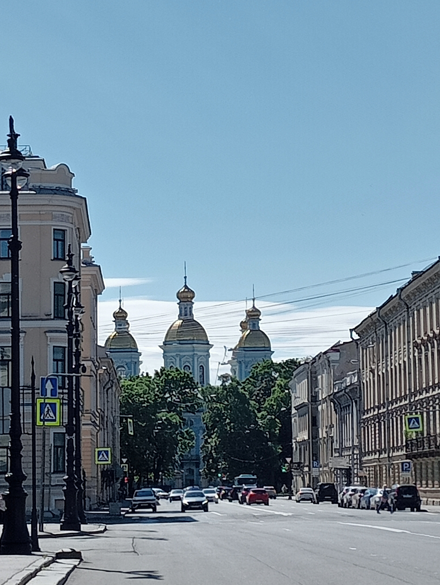 The image captures a street scene in Saint Petersburg, Russia, on a bright, clear day. The perspective is from a street level, looking down a wide avenue lined with classical architecture. In the distance, three gilded onion domes of a prominent church, likely St. Nicholas Naval Cathedral, rise above a dense canopy of green trees. The buildings on either side of the street exhibit ornate facades with multiple windows and decorative elements.
On the left side, a tall, dark lamppost with intricate detailing stands prominently in the foreground. Traffic signs, including a pedestrian crossing sign with a blue background and a white silhouette of a person walking, are visible on this side of the street. The street itself is paved with asphalt and marked with white lane lines.
Several cars are driving on the road, with some parked along the right side. The traffic appears moderate. The sky is a clear, pale blue, with a few wispy white clouds visible in the distance. The overall atmosphere is one of an active urban environment on a sunny day.