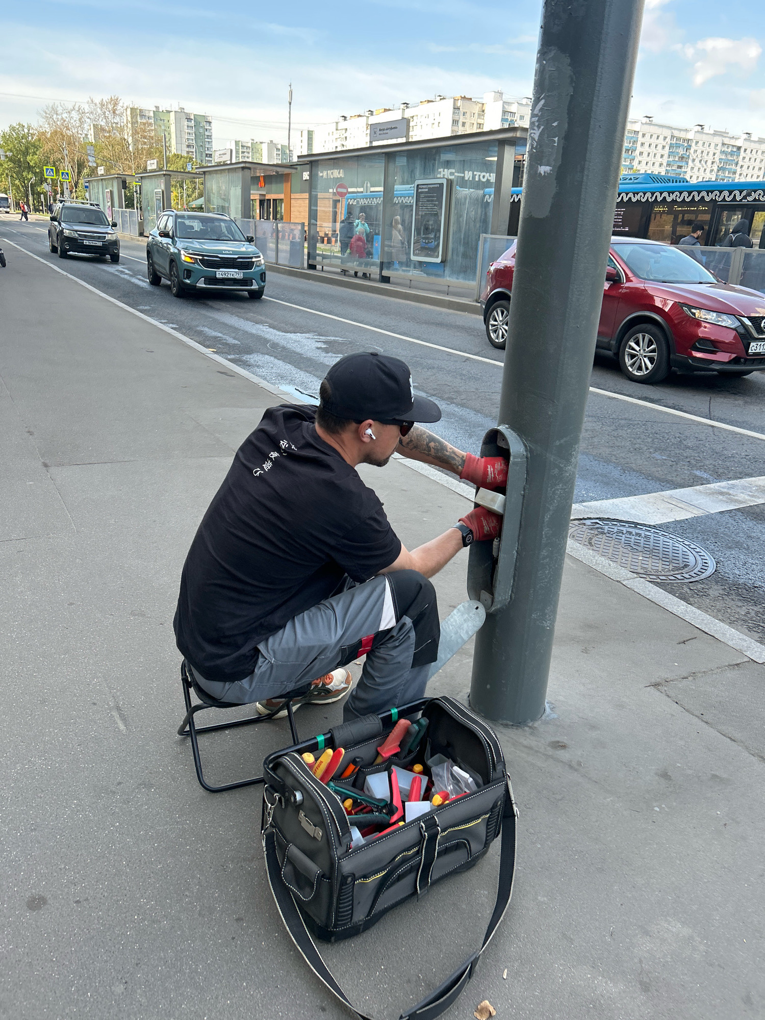 A man wearing a black t-shirt, grey pants, and red gloves is working on a gray pole on a sidewalk in Moscow, Russia. He is seated on a small folding stool with a toolbox in front of him. The toolbox contains various tools, including screwdrivers and pliers. In the background, there is a street with cars and a bus stop. The sky is overcast.