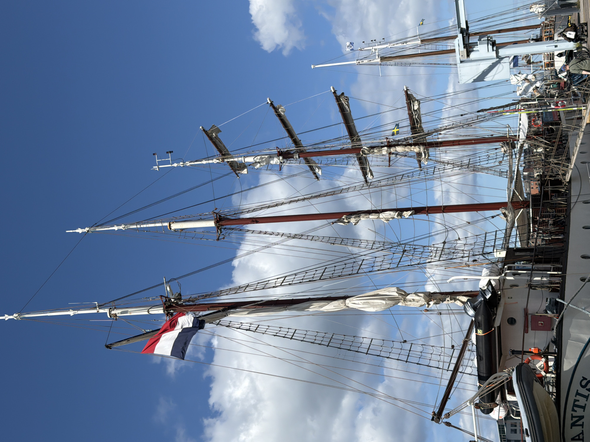 This is a medium shot, eye-level, vertical, exterior photograph of the tall ship "Atlantis" docked in the Western Harbour (Västra Hamnen) of Malmö, Sweden, on a clear, partly cloudy day. The ship has multiple masts, with sails furled and secured, and is adorned with rigging and lines. A Dutch flag is prominently displayed on one of the masts. The hull of the ship is white, with the name "ATLANTIS" visible near the bow. Several smaller inflatable boats are stowed on deck, along with life preservers and other nautical equipment. In the background, other docked vessels and the modern architecture of the Västra Hamnen district are visible. Two individuals are present near the stern of the ship, one walking with a bag and the other seated in a mobility scooter. The sky is a vibrant blue with scattered white clouds.