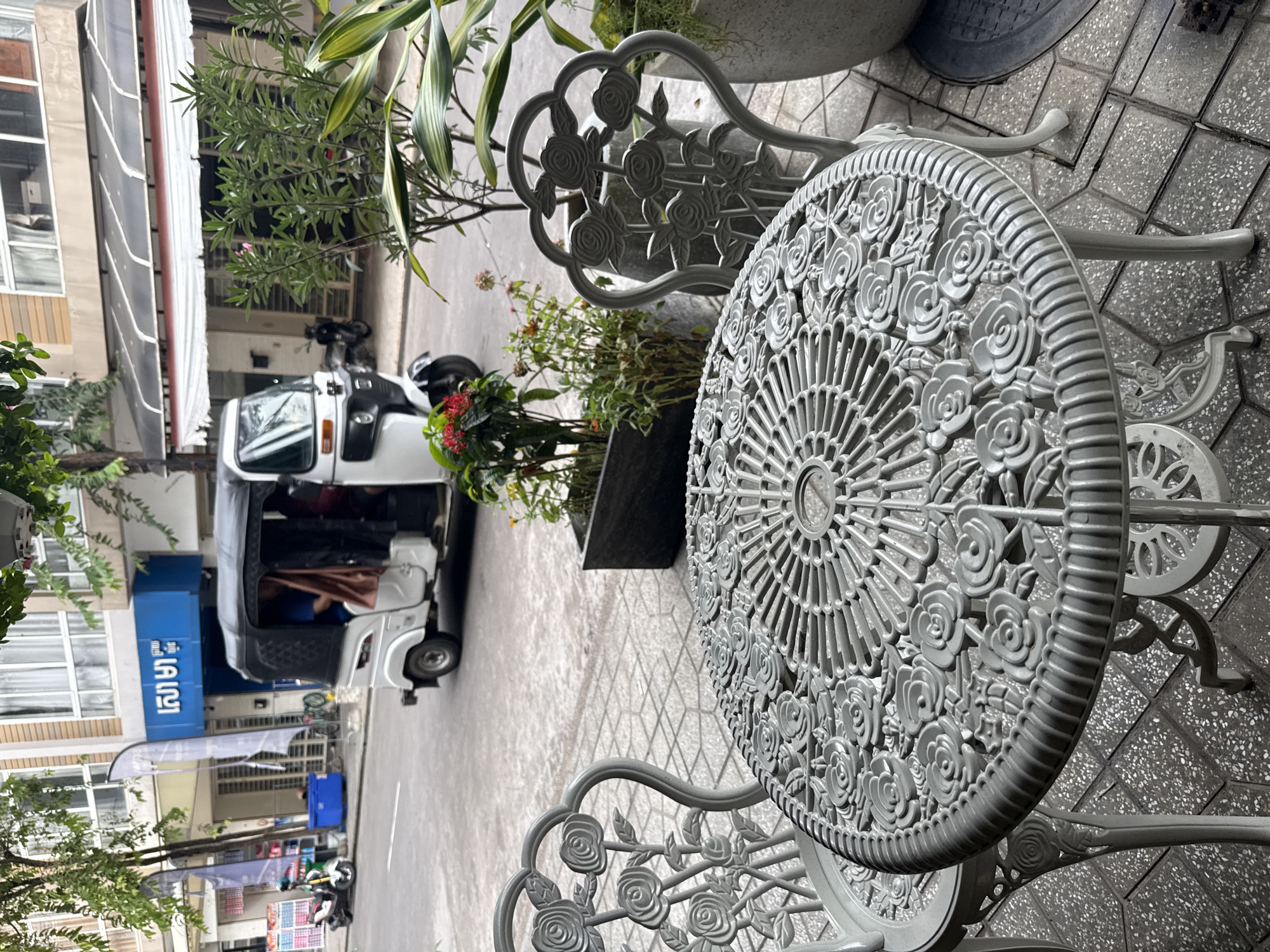 This is a medium shot of a sidewalk cafe scene in Khan Chbar Ampov, Cambodia. In the foreground, a white wrought-iron table with an ornate floral design is set with two matching chairs. The table is round and features a detailed radial pattern of roses and leaves. The chairs have curved backs adorned with similar rose motifs. The sidewalk is paved with grey interlocking tiles.
Beyond the cafe furniture, a street is visible with a white tuk-tuk driving away from the camera. The tuk-tuk has a black canopy and two figures are seated inside. The street appears to be paved with asphalt. On the left side of the street, there is a blue awning over a shop with a blue sign featuring white Khmer script. Several parked motorbikes and a scooter are also visible in front of the shop. To the right of the tuk-tuk, a large potted plant with green and yellow striped leaves is situated next to a large concrete planter containing smaller plants with red and green foliage. The background consists of a multi-story building with windows and awnings. The overall impression is a street scene in a Southeast Asian city, likely during daytime.