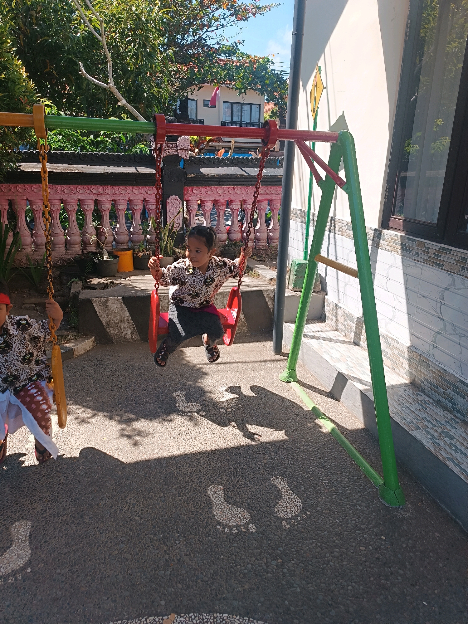 In Tabanan, Indonesia, two young children are playing on a swing set in a courtyard. The swing set is constructed with green and red metal poles. The child on the right, wearing a patterned shirt and dark pants, is actively swinging forward. The child on the left, partially visible, is also on a swing. The ground is a textured gray surface, possibly asphalt or concrete, with small pebbles embedded in it. In the background, a pink ornamental fence with white posts separates the courtyard from a building. Behind the fence, there are some potted plants and trees. The overall scene is illuminated by bright sunlight, casting shadows on the ground. The building in the background appears to be a house or a low-rise structure with a light-colored wall and a dark-framed window.