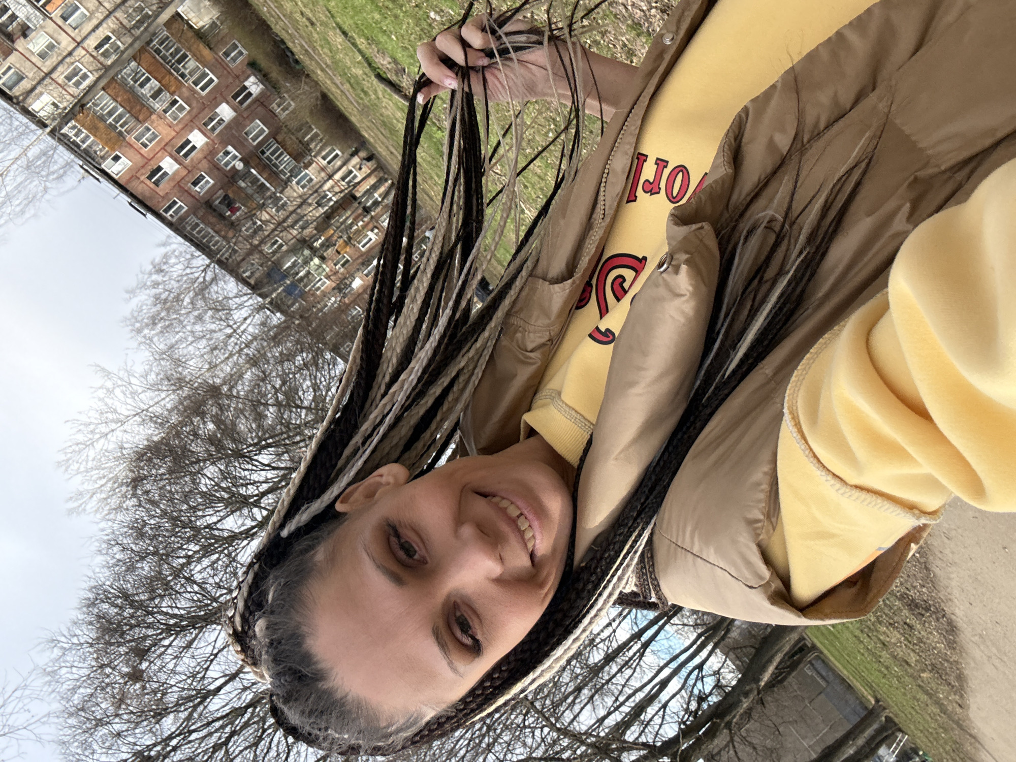 A woman with long brown and blond braids is smiling at the camera. She is wearing a yellow sweatshirt and a beige vest. In the background is a multi-story brick building and trees with bare branches. The image was taken in Fedorino, Russia.